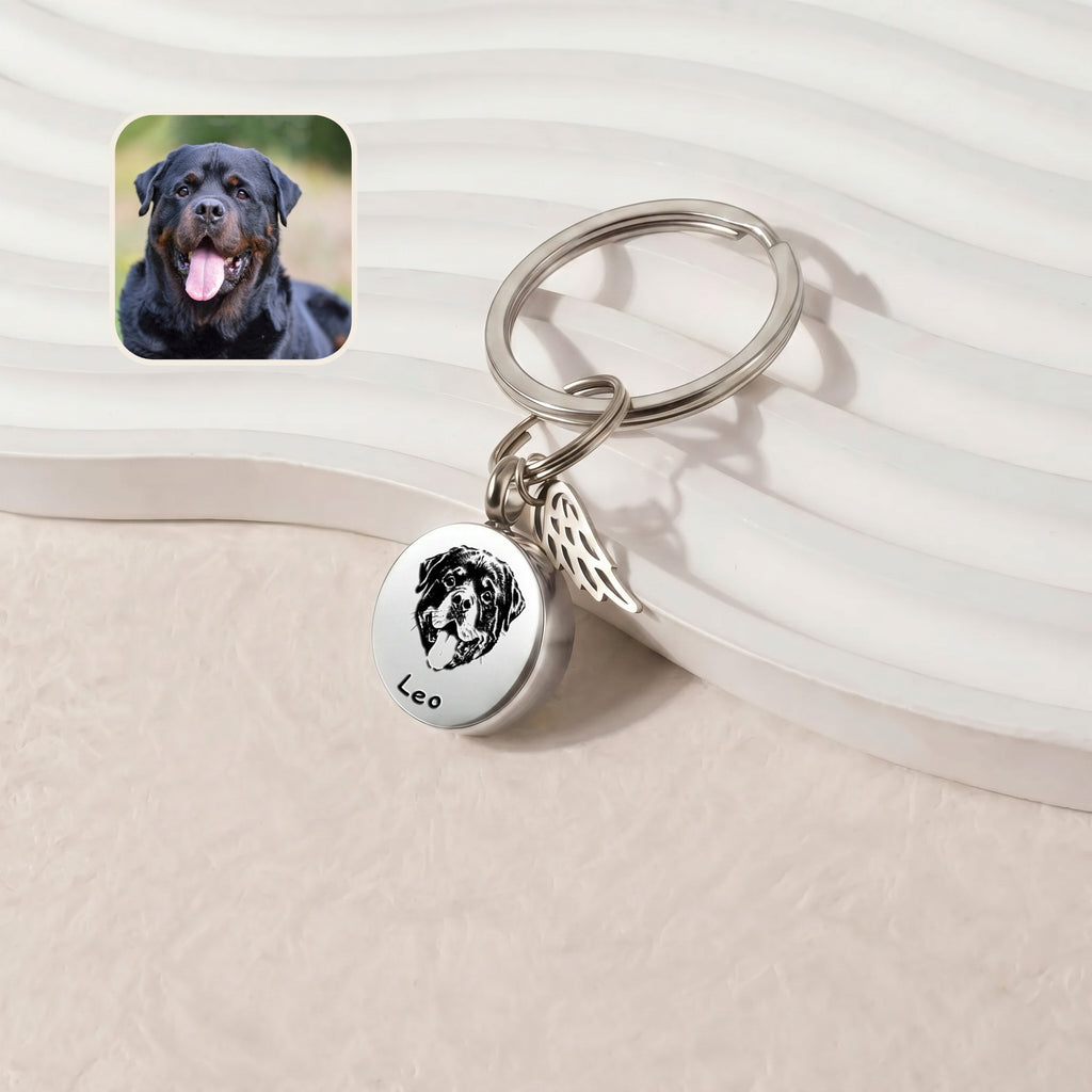 Rhodium-plated pet cremation disc keychain &mdash; hand-drawn portrait engraved on a 20mm screw-top disc with permanently-attached angel-wing memorial charm, leaned against a ribbed ceramic tray on a cream plush surface beside a reference dog photo inset
