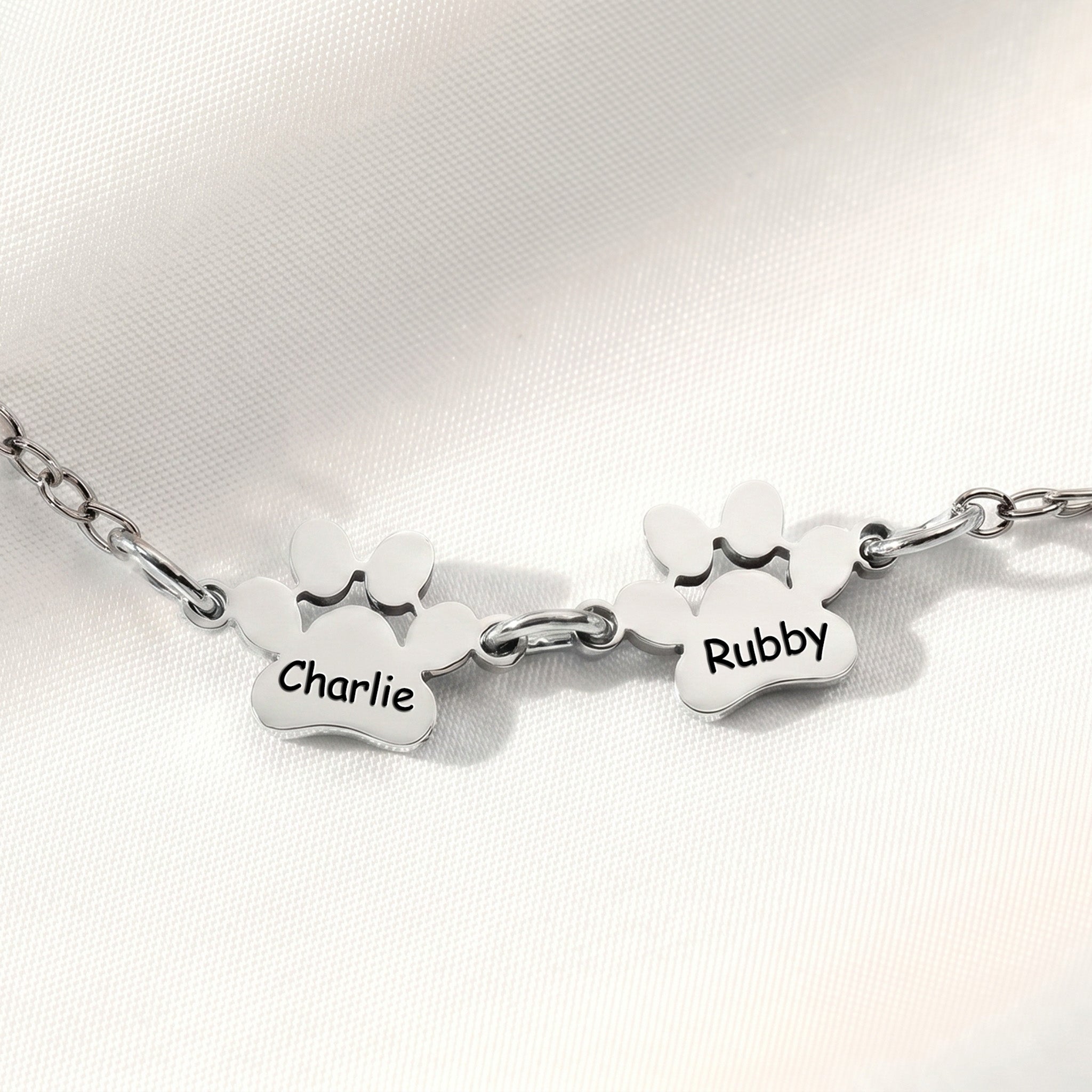 Two rhodium paw name charms on an adjustable chain bracelet on white satin