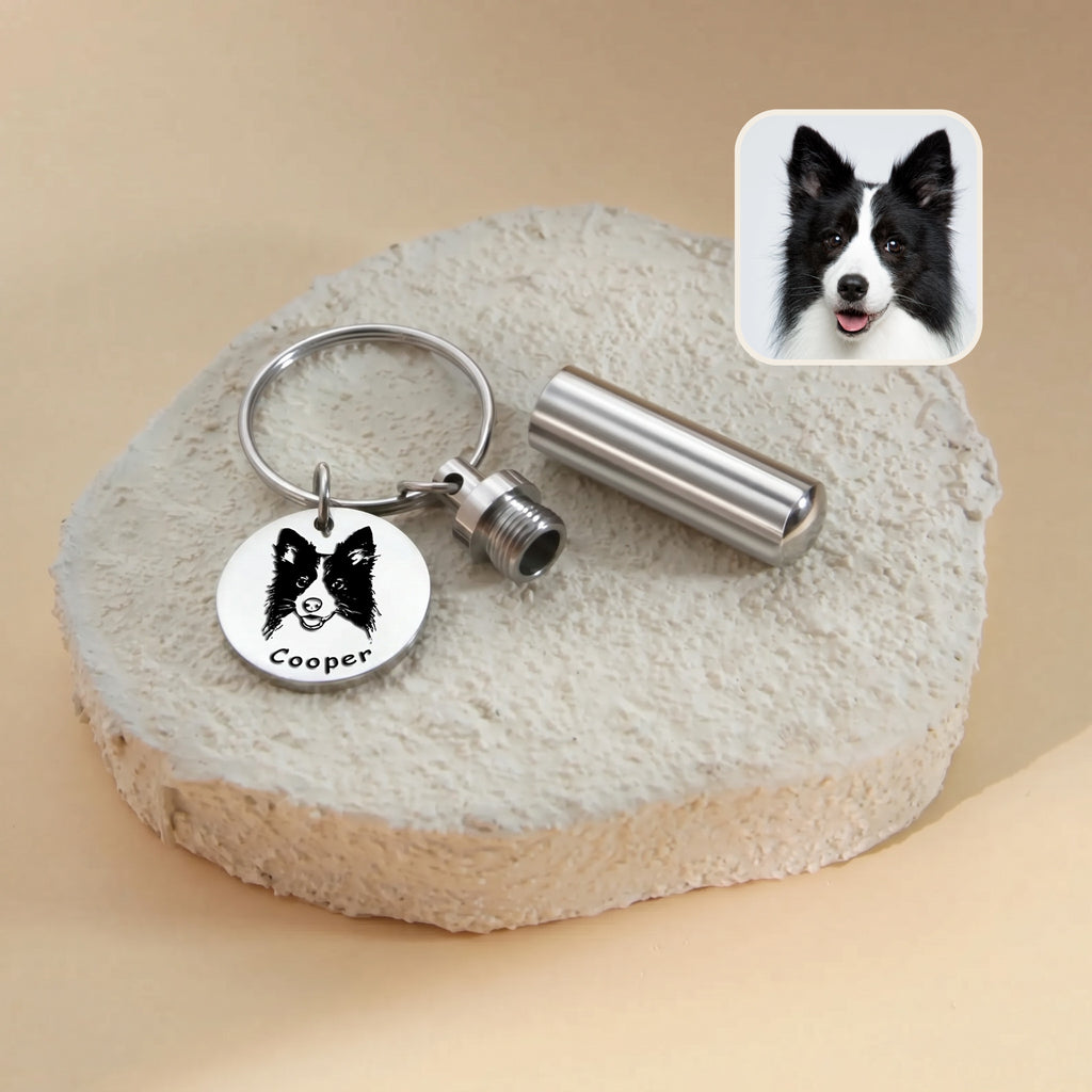 Rhodium-plated pet portrait and ashes cylinder keychain close-up &mdash; engraved disc with hand-drawn border collie portrait and the name Cooper beside a screw-cap cylinder urn, with a reference photo inset on a textured stone surface