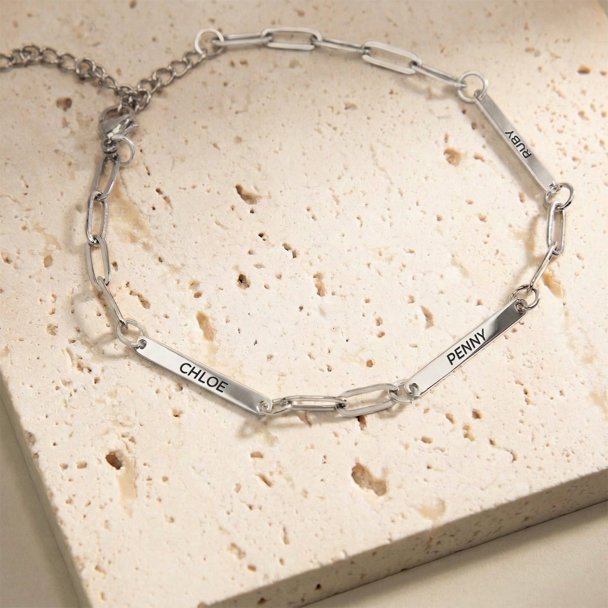 Silver rhodium paperclip chain pet name bracelet with three engraved name bars on a cream stone block