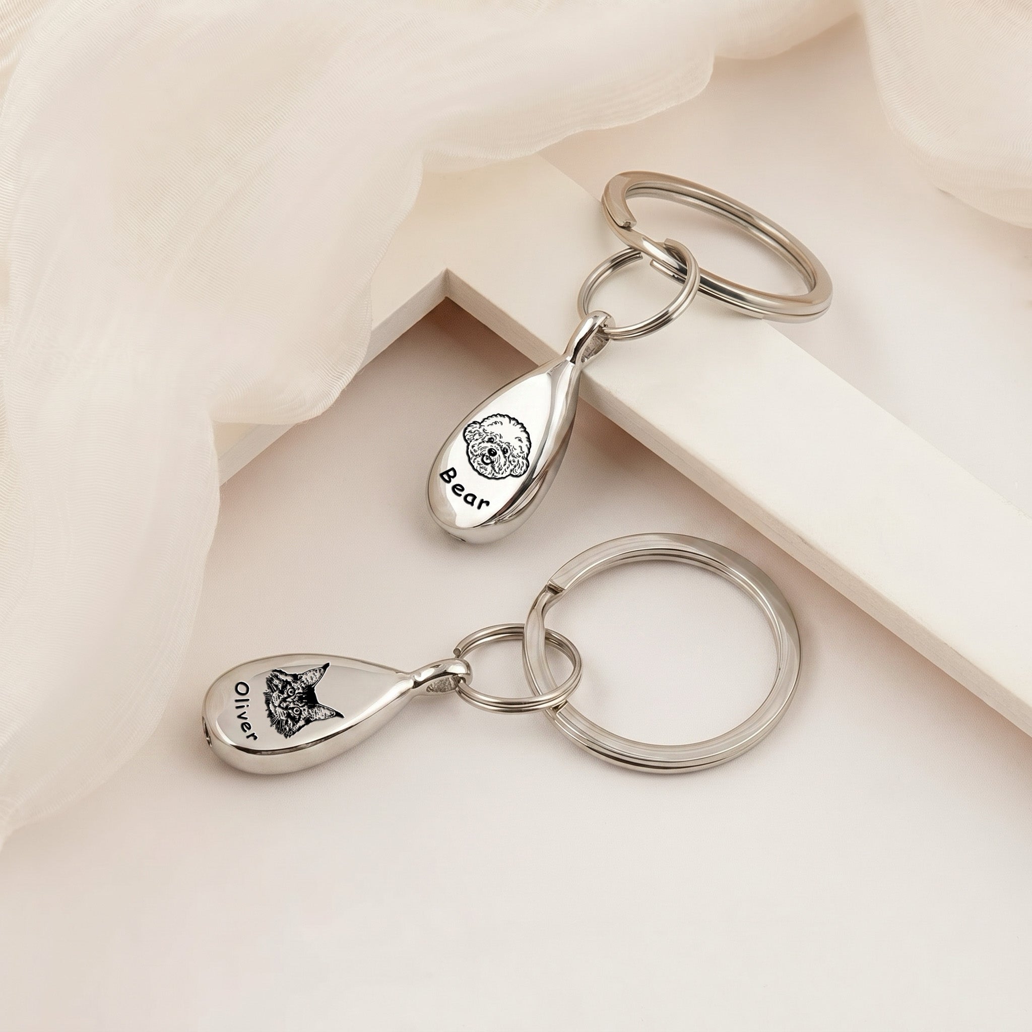 Pair of rhodium-plated pet portrait ashes teardrop keychains arranged on a cream plinth under soft gauzy fabric &mdash; both pendants engraved with hand-drawn pet portraits and names