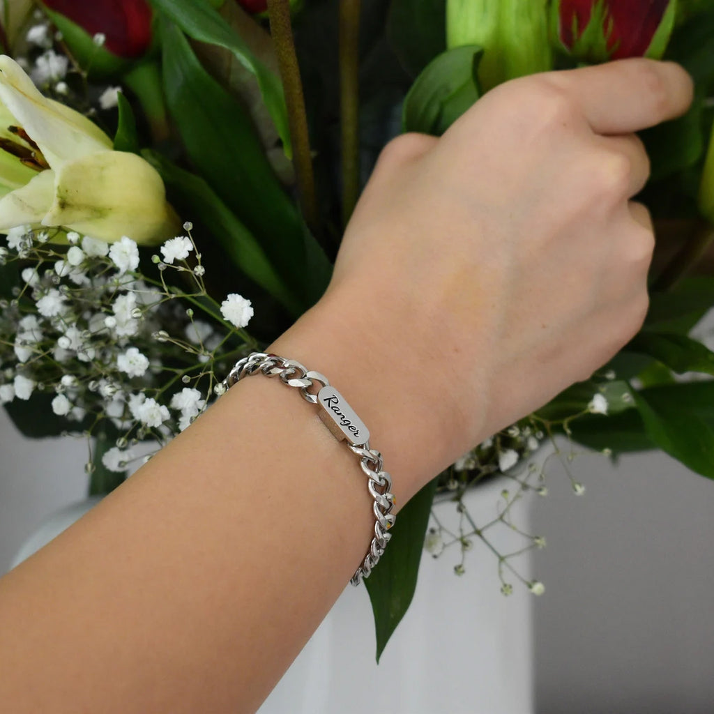 Rhodium silver pet name bracelet with a script-engraved nameplate, worn on the wrist beside flowers