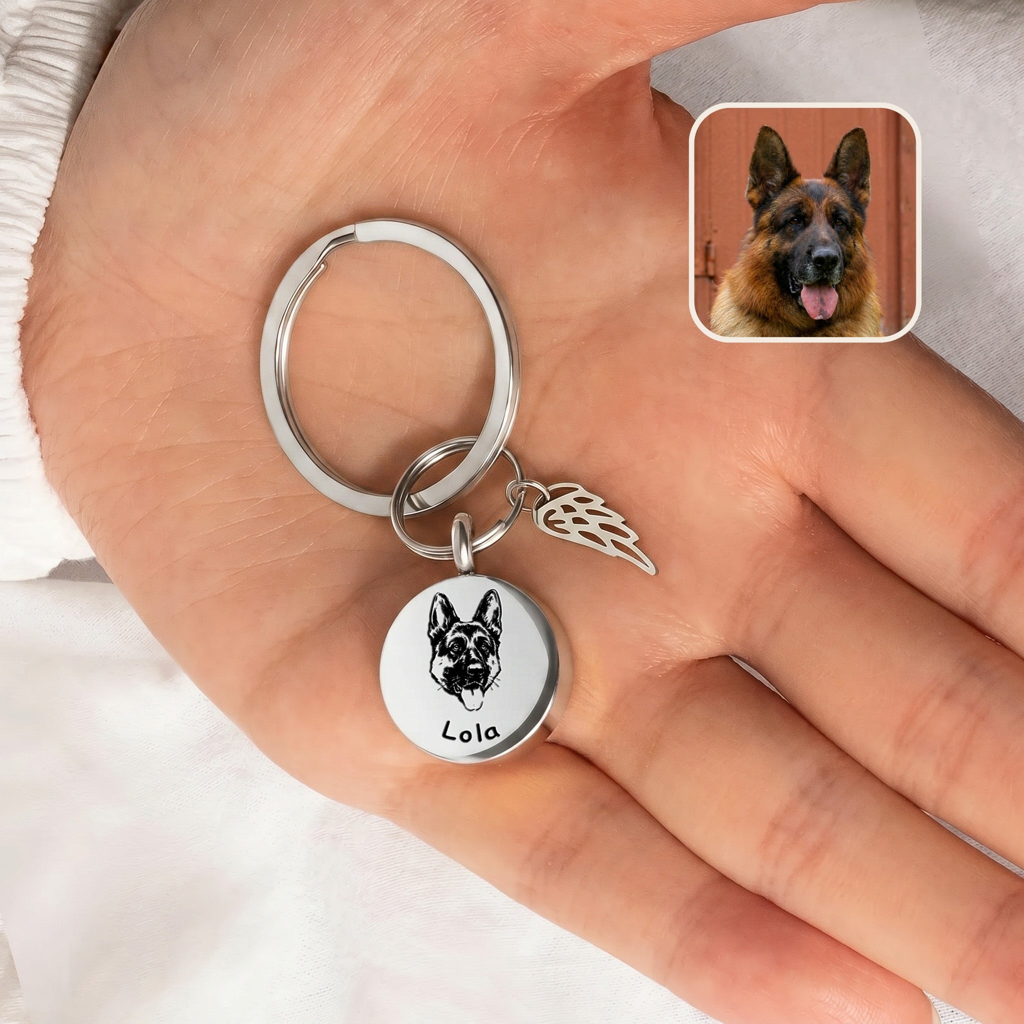 Rhodium-plated pet cremation disc keychain with angel-wing memorial charm shown in an open hand beside a reference dog photo inset, scale-only no in-use carry