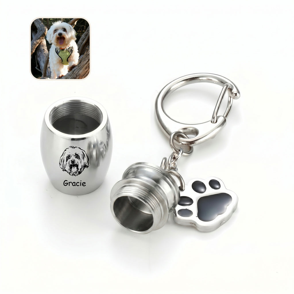 Rhodium pet urn keychain shown with the screw cap opened to reveal the inner chamber, paw-print side charm alongside the capsule, a small dog reference photo inset