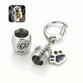 Rhodium pet urn keychain shown with the screw cap opened to reveal the inner chamber, paw-print side charm alongside the capsule, a small dog reference photo inset
