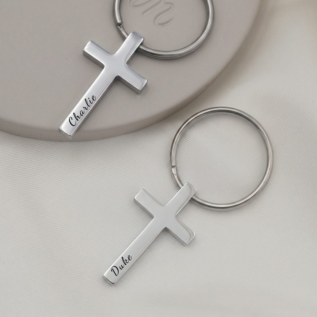 Rhodium pet cross keychain pair shown on ceramic tray with soft white fabric — front-face hand-drawn portrait + pet name engraving visible