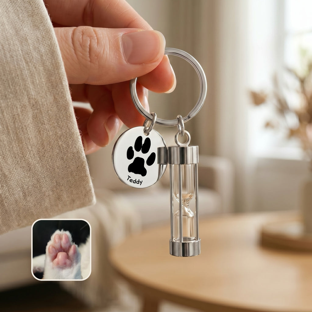 Rhodium-plated pet memorial hourglass keychain held in hand showing paw-print engraving option on disc and glass hourglass urn, with cat paw reference photo