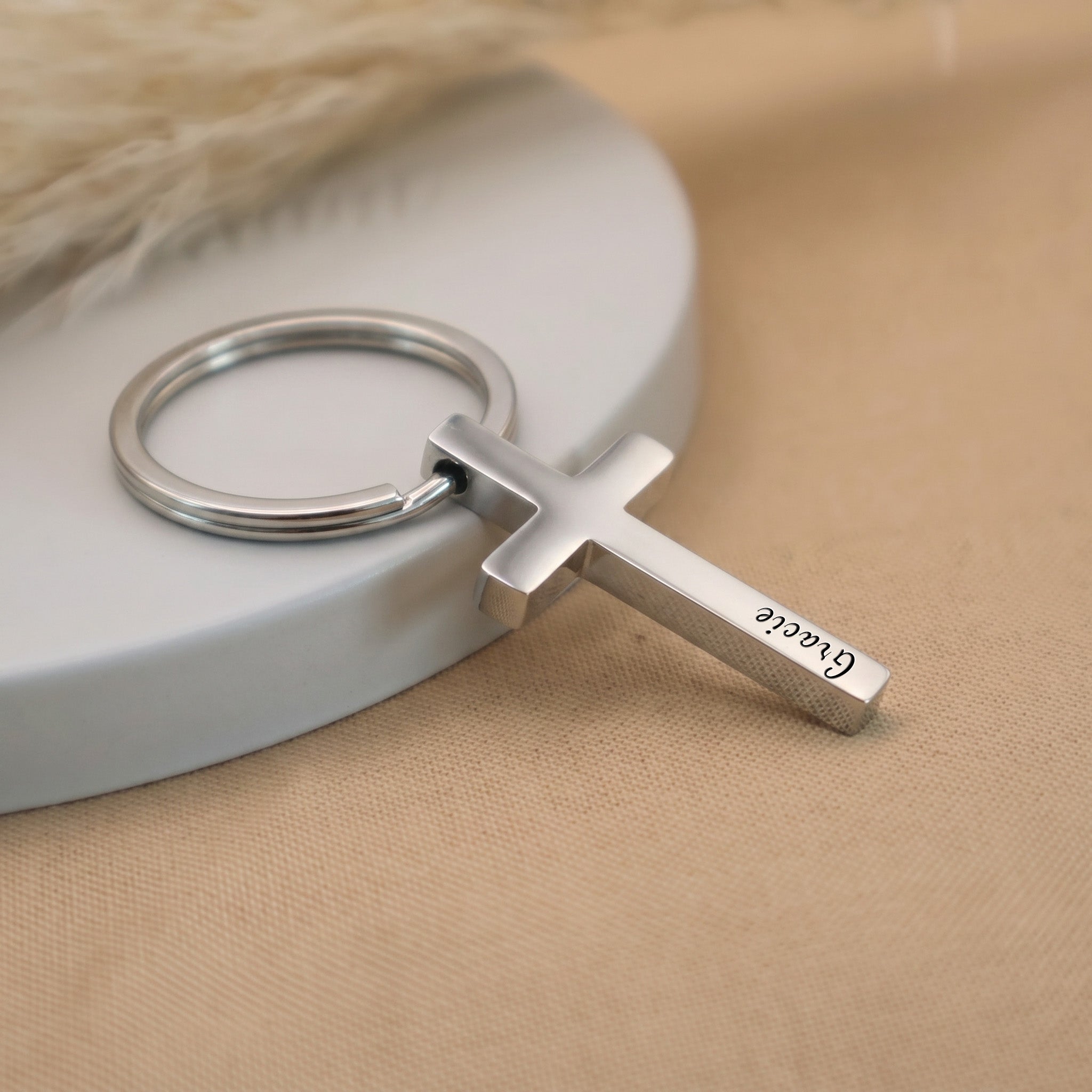 Rhodium pet cross keychain angled on a ceramic tray with beige fabric — full pendant and 25mm key ring visible in everyday-carry scene