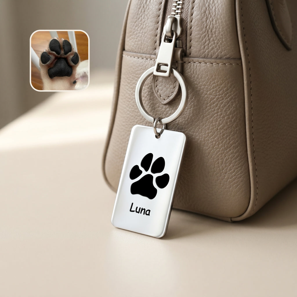 Rhodium plate keychain clipped to a taupe handbag zipper, paw print motif and name 'Luna' engraved on the rectangular plate