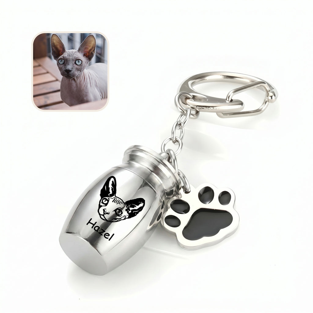 Angled rhodium pet urn keychain close-up on a clean white surface with a cat reference photo inset, showing the hand-drawn portrait engraving on the capsule face