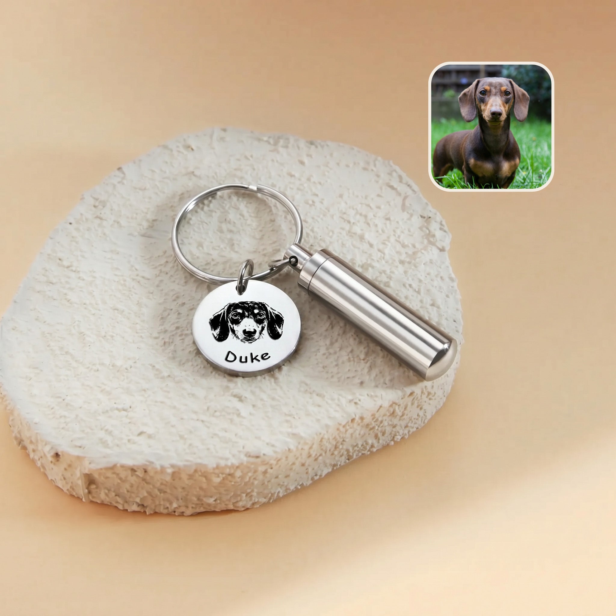 Rhodium-plated pet portrait and ashes cylinder keychain close-up &mdash; engraved disc with hand-drawn dachshund portrait and the name Duke beside the cylinder urn, with a reference photo inset
