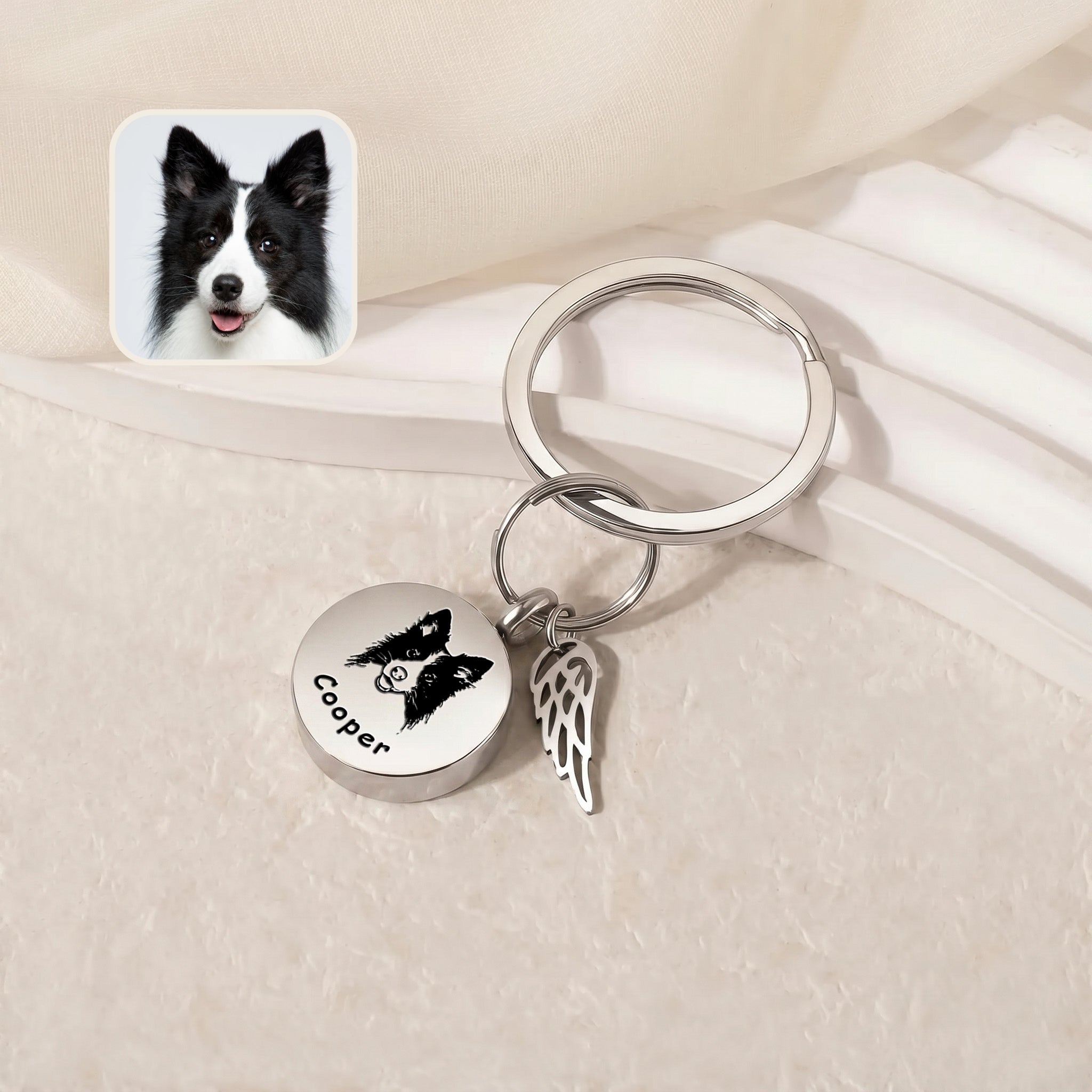 Rhodium-plated pet cremation disc keychain &mdash; engraved disc and angel-wing memorial charm on a pale stepped tray beside a black-and-white reference pet photo inset
