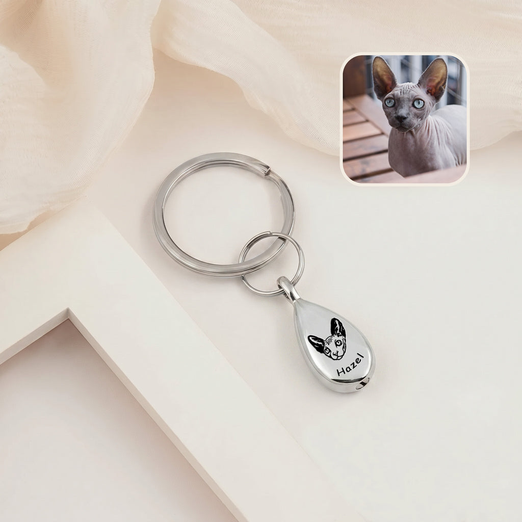 Rhodium-plated pet portrait ashes teardrop keychain on a cream ledge with sheer fabric &mdash; engraved with a hand-drawn cat portrait and name, with a cat reference photo inset