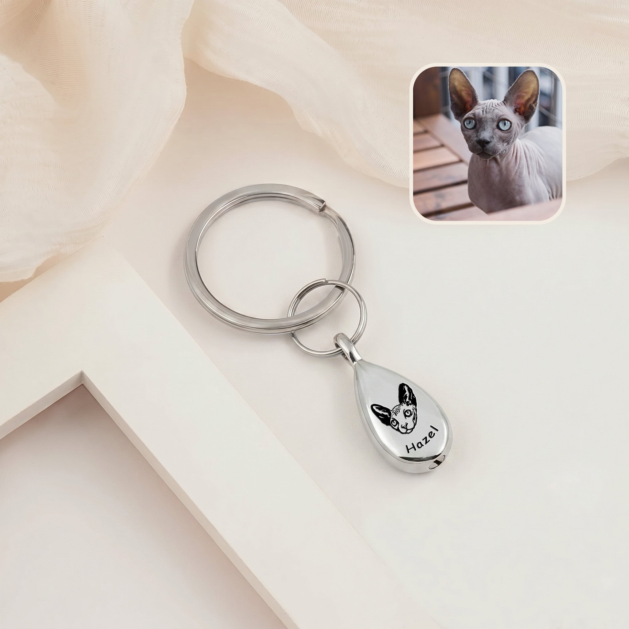 Rhodium-plated pet portrait ashes teardrop keychain on a cream ledge with sheer fabric &mdash; engraved with a hand-drawn cat portrait and name, with a cat reference photo inset