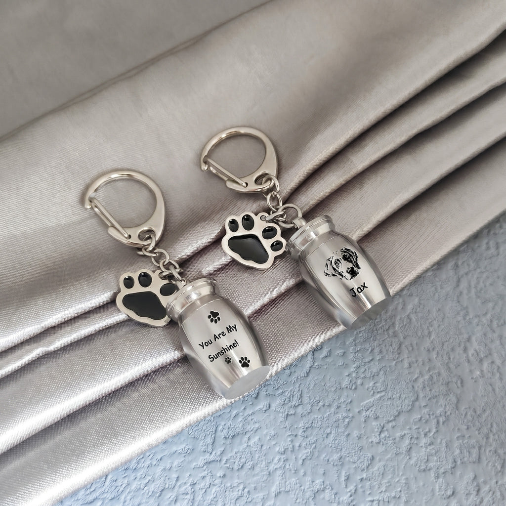 Two rhodium pet urn keychains arranged on draped satin fabric, one engraved with a verse and name, the other engraved with a hand-drawn portrait and name