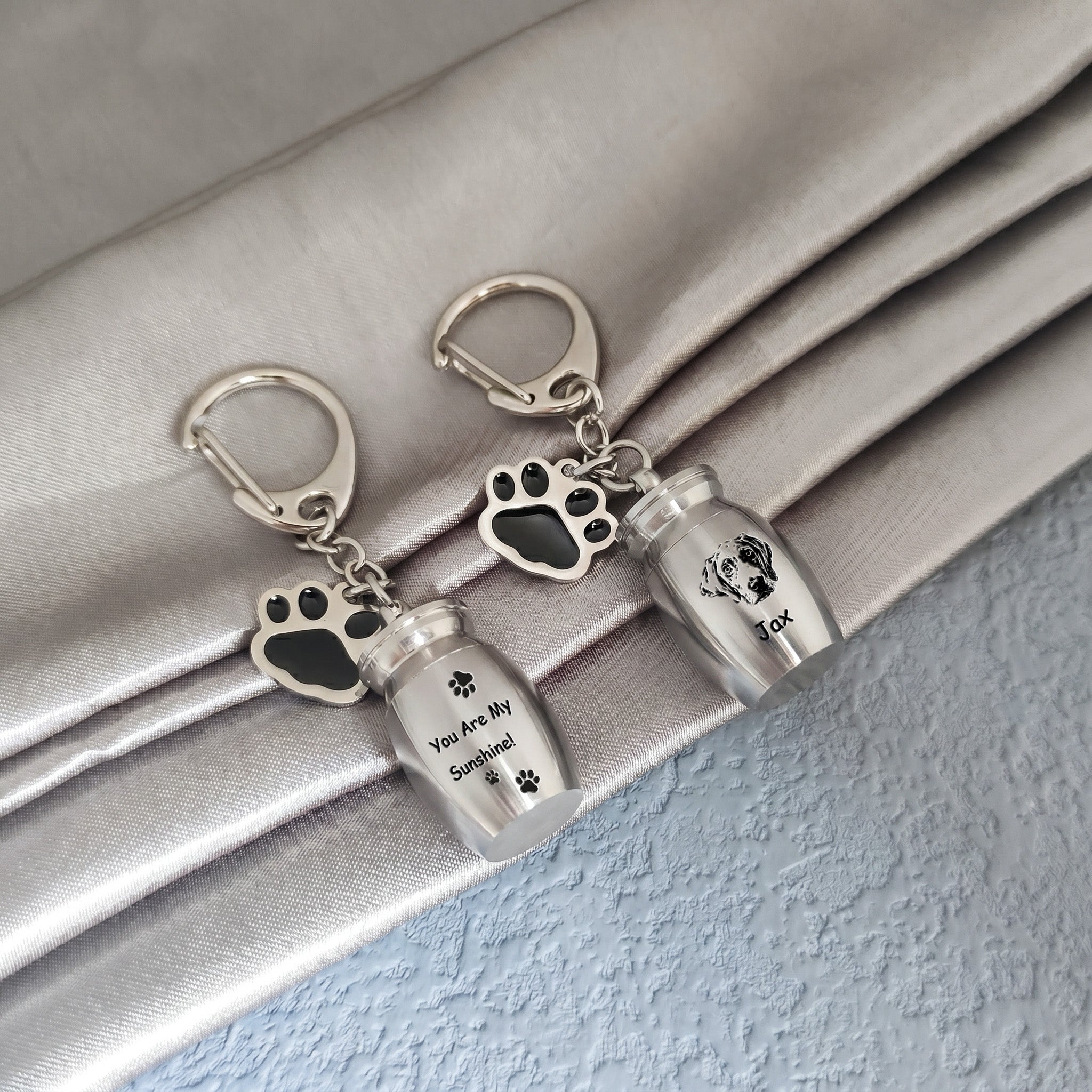 Two rhodium pet urn keychains arranged on draped satin fabric, one engraved with a verse and name, the other engraved with a hand-drawn portrait and name