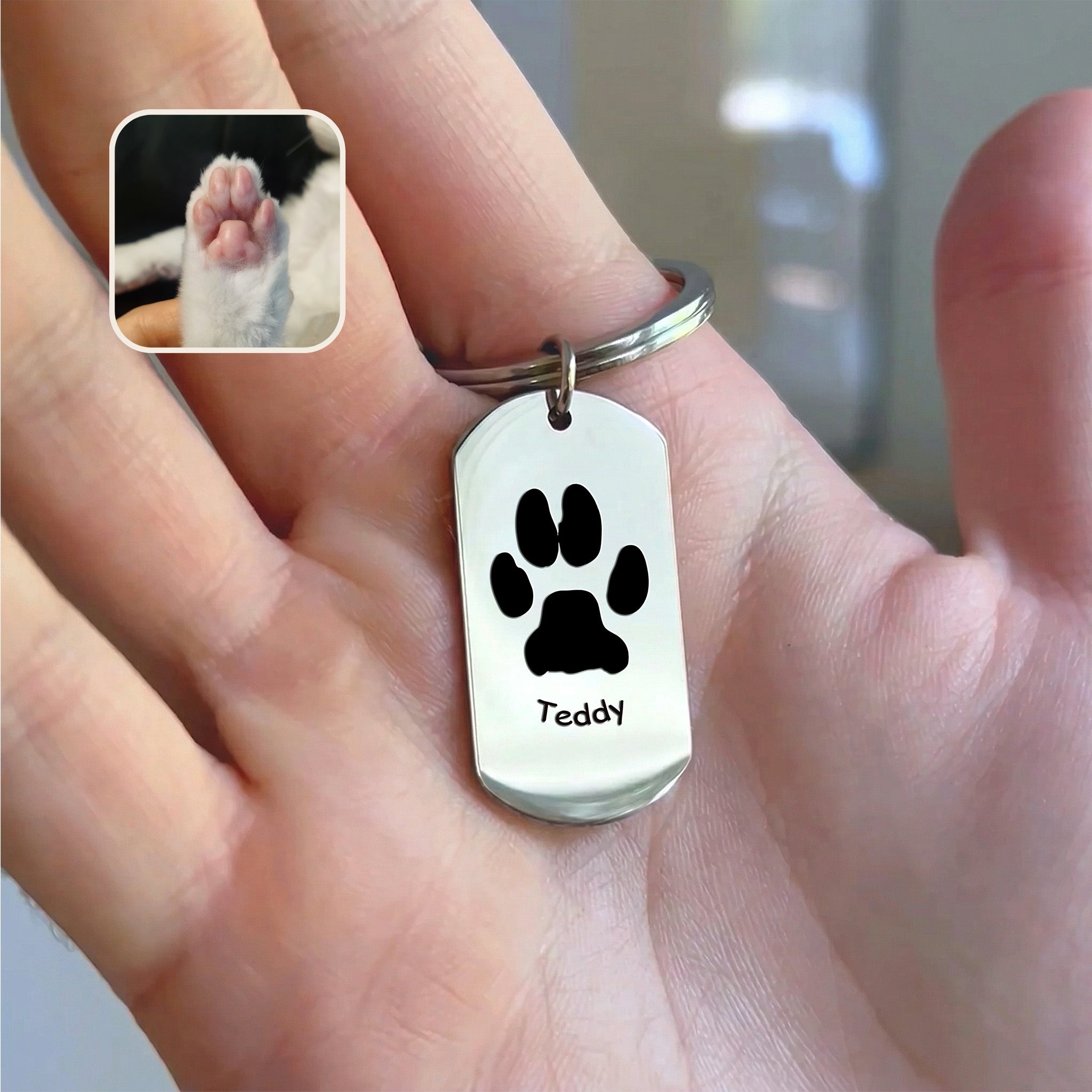 Rhodium plated dog tag keychain with paw-print motif and pet name engraving, held in hand beside paw reference photo, warm indoor styled scene