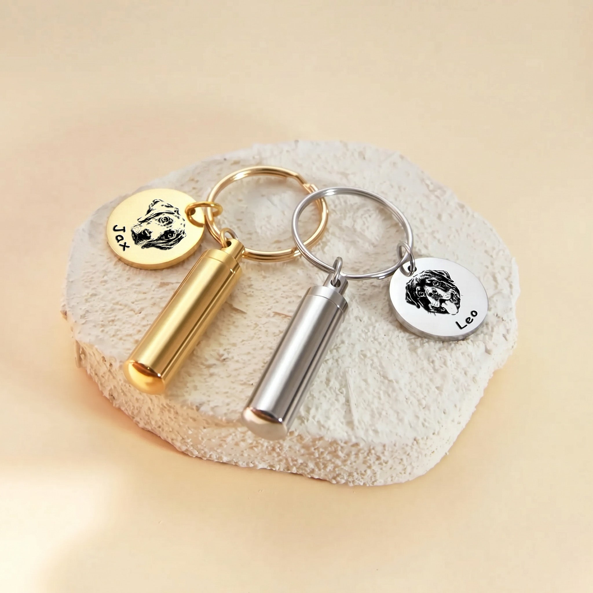 Pet portrait and ashes cylinder keychains shown in both finishes &mdash; 18k gold and rhodium silver paired on a textured stone surface