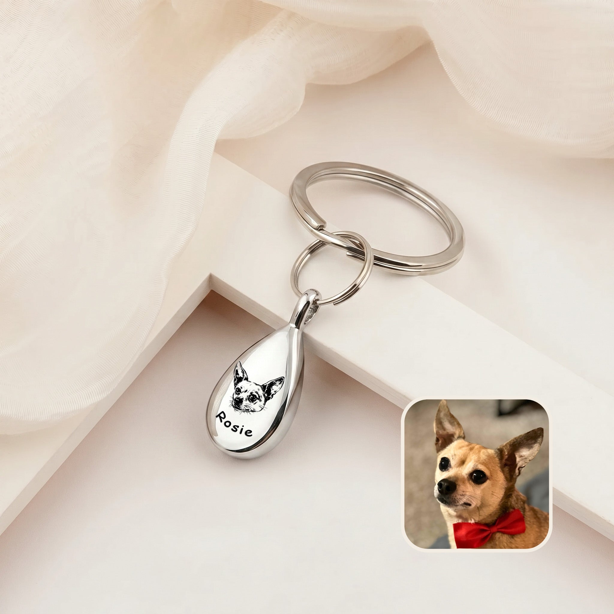 Rhodium-plated pet portrait ashes teardrop keychain on a cream ledge with sheer fabric &mdash; engraved with a hand-drawn dog portrait and name, with a dog reference photo inset