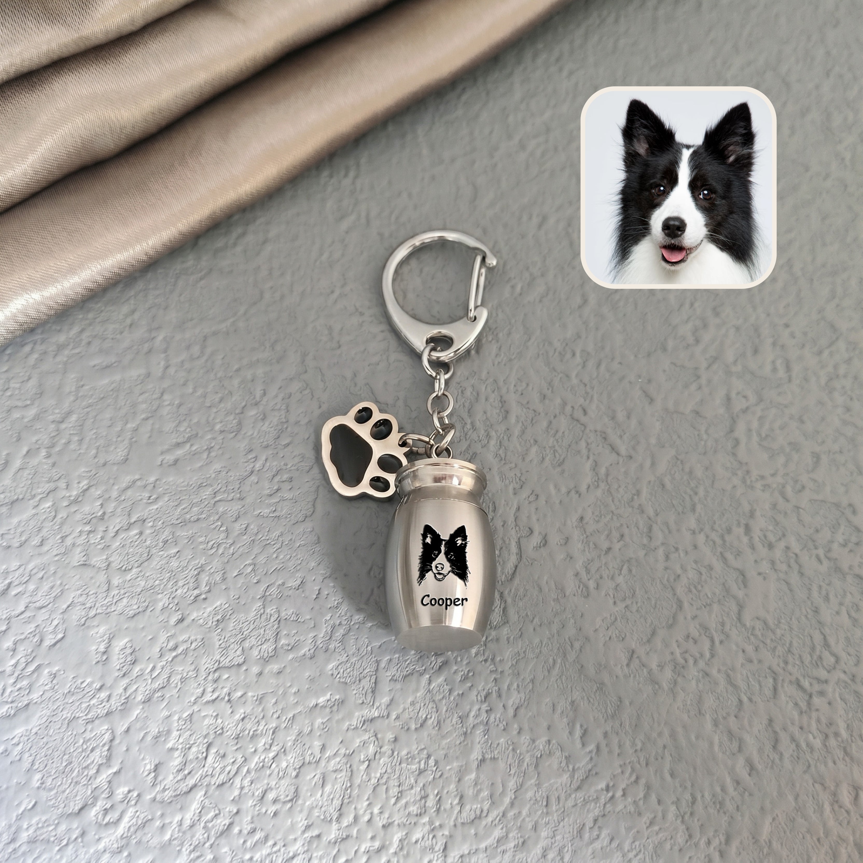 Rhodium pet urn keychain staged against a textured wall background with a dog reference photo inset, hand-drawn portrait engraved on the capsule face