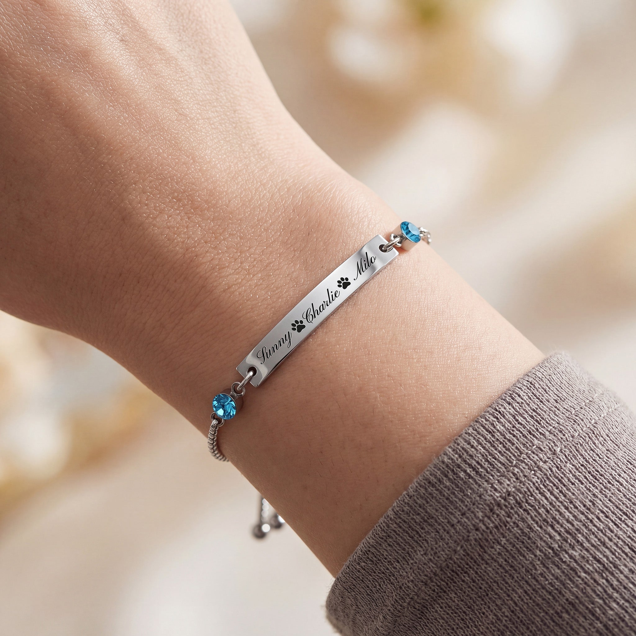 Rhodium name bar bracelet on a wrist, three engraved names with paw print separators and blue birthstones, slider chain