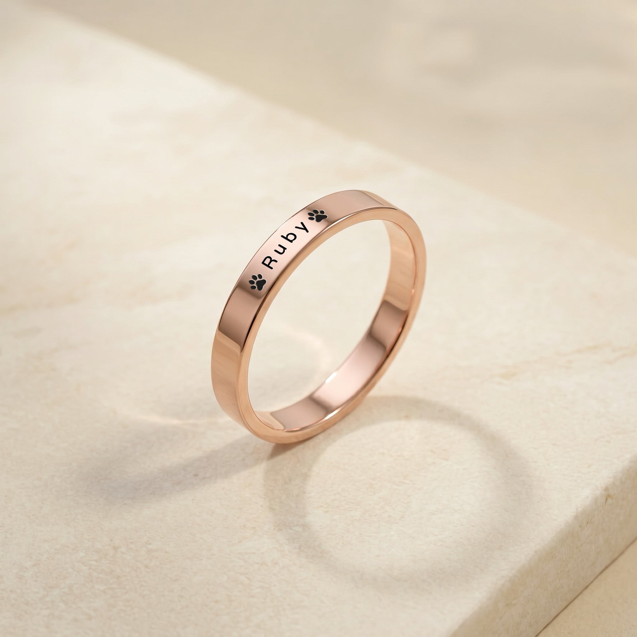 Rose gold pet name band ring engraved with a pet’s name and a paw, angled on a cream stone plinth