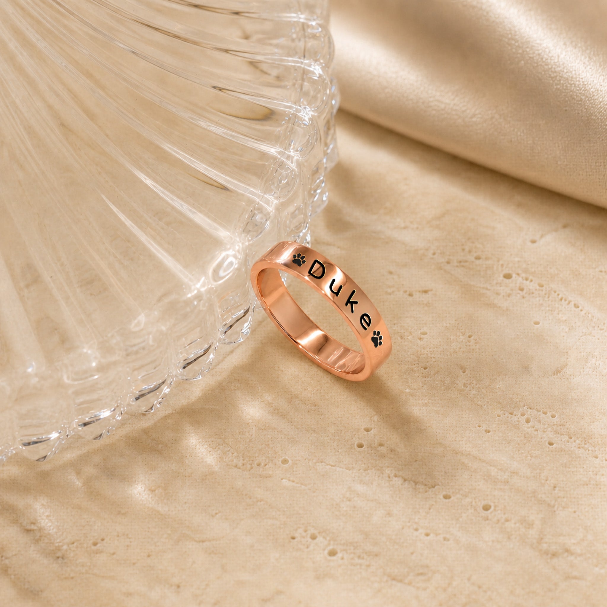 18K rose gold-plated pet name stacking ring engraved with a name and paw prints, beside a fluted glass dish on travertine
