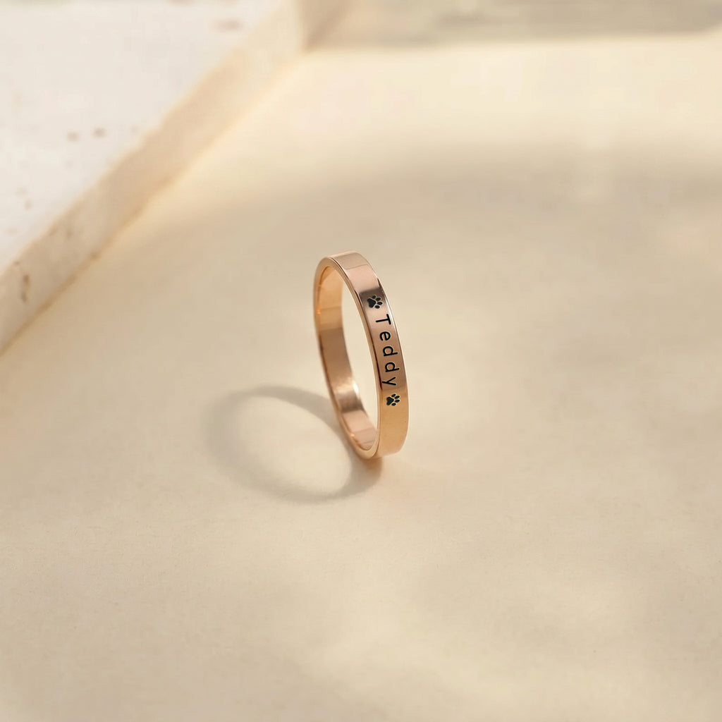 Rose gold pet name band ring standing on a warm stone surface, name and paw engraving facing forward