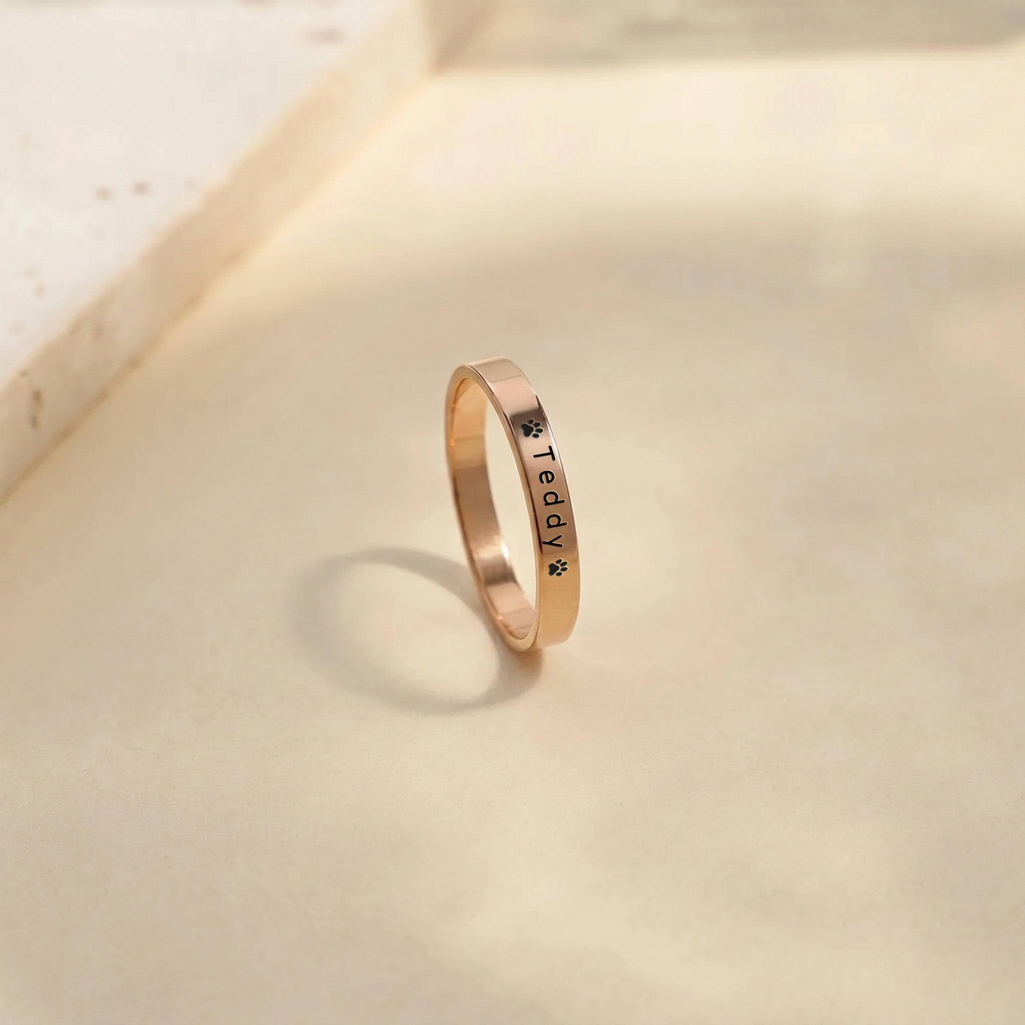 Rose gold pet name band ring standing on a warm stone surface, name and paw engraving facing forward