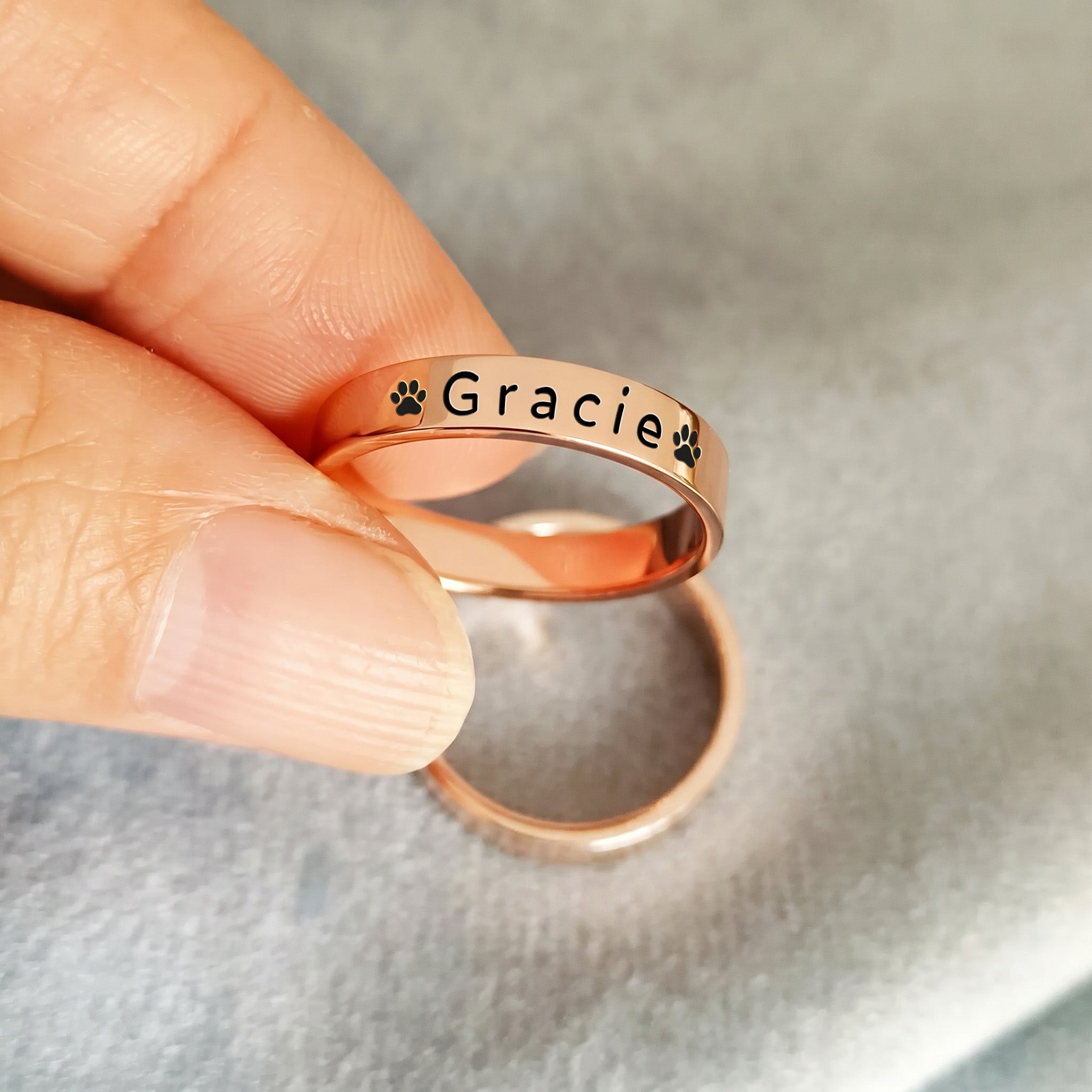 18K rose gold-plated pet name stacking ring held between fingertips against a soft grey background