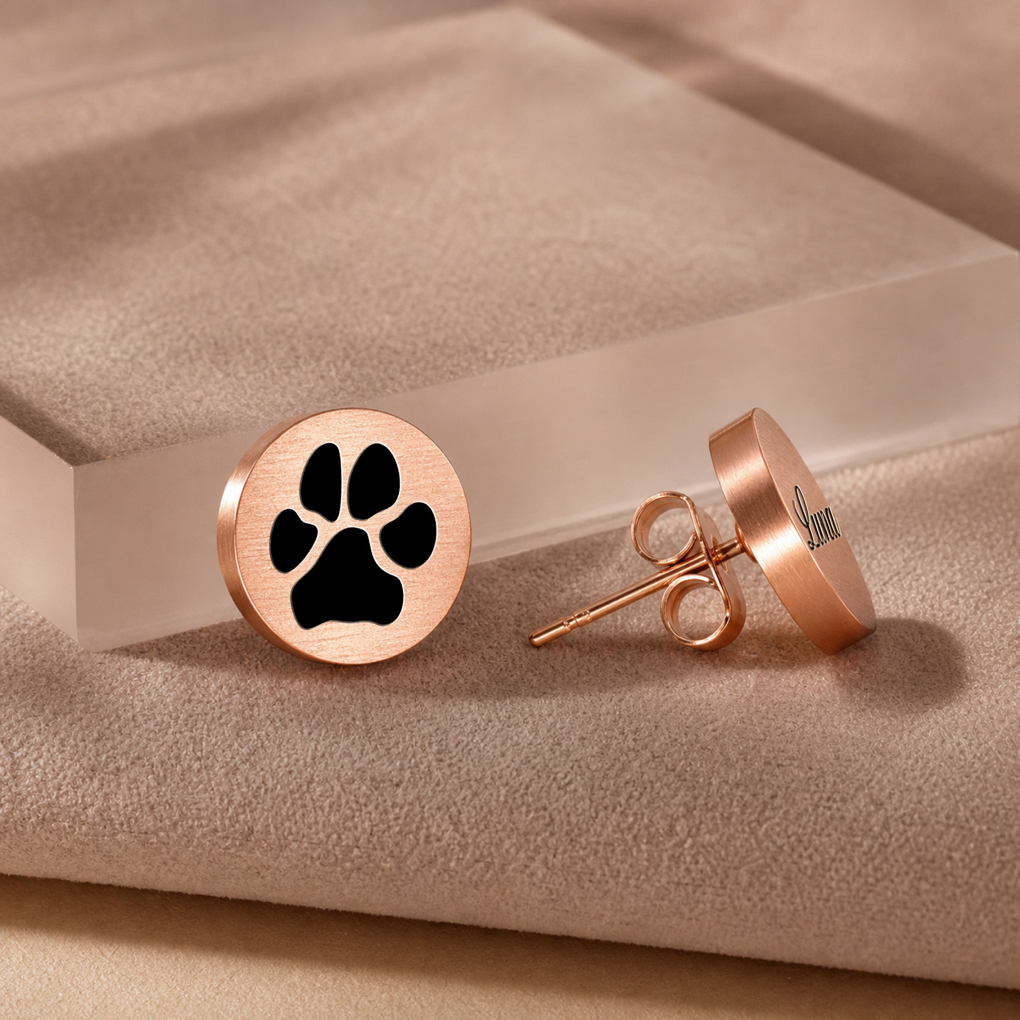 Rose gold pet name stud earring with script-engraved pet name on a round disc, on a beige textured block