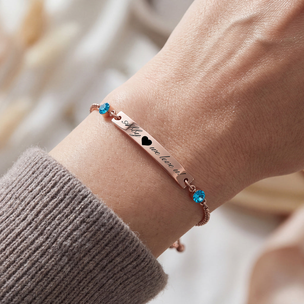 18K rose gold name bar bracelet worn on a wrist, two engraved names with a heart and blue birthstones at each end on a slider chain