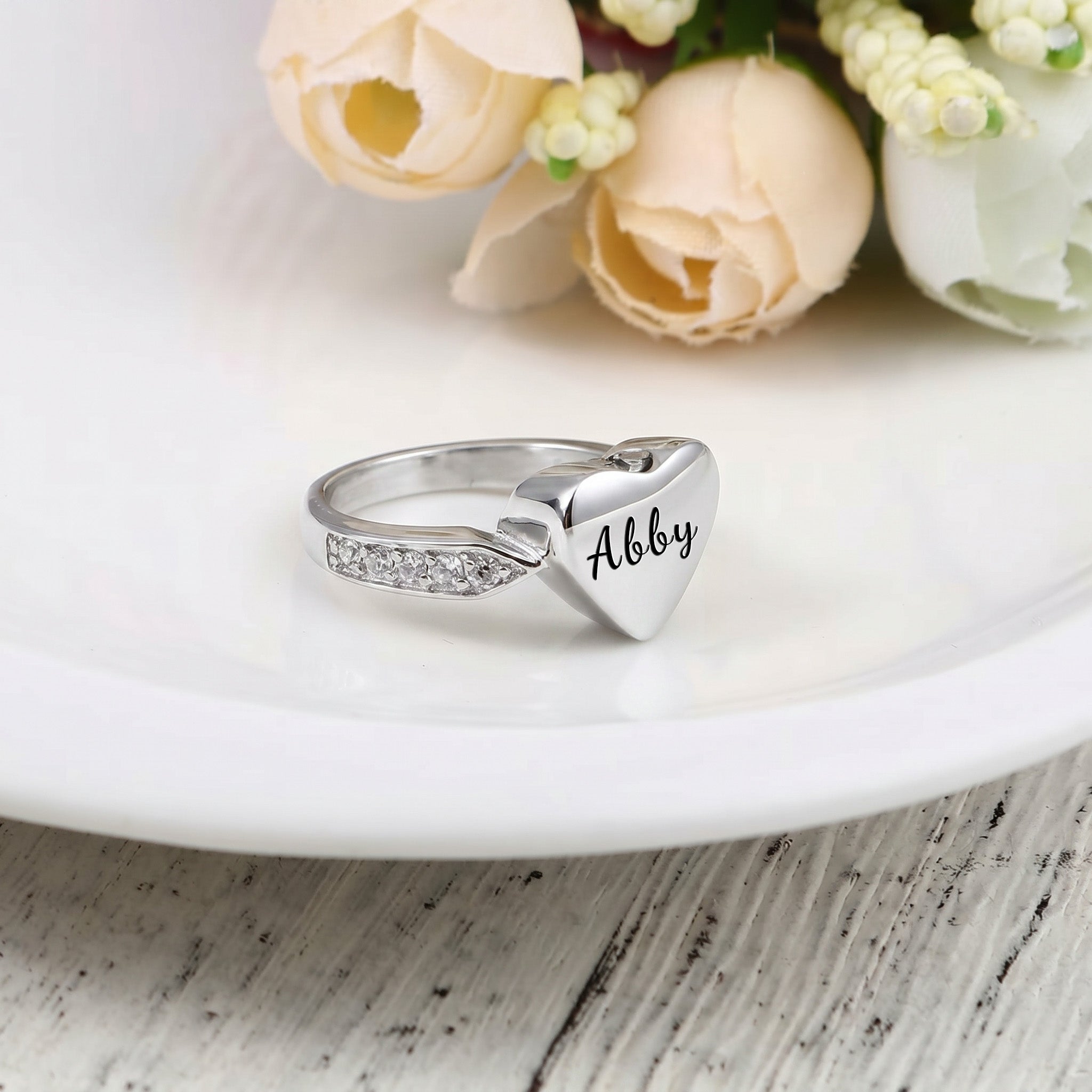 Silver heart urn ring with pet name engraved in script, on a white ceramic plate with roses