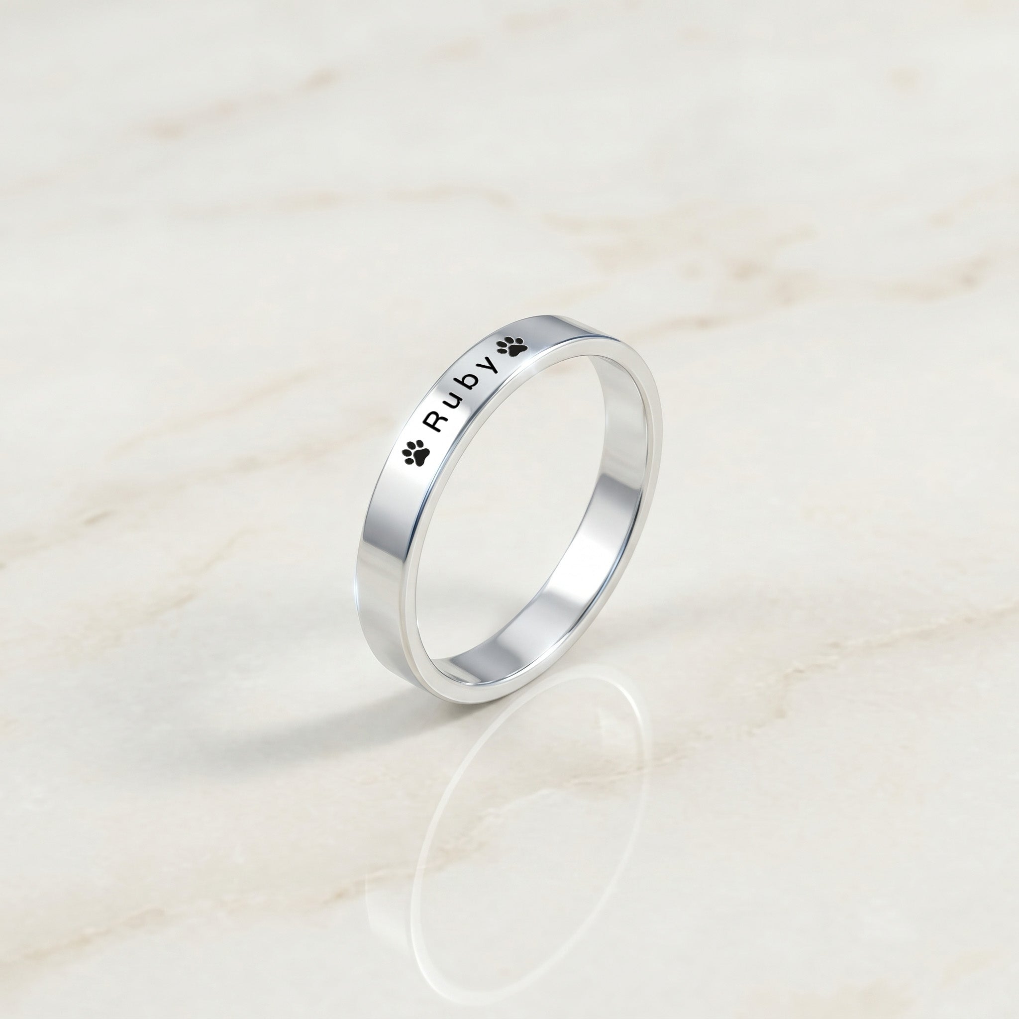 Silver pet name band ring engraved with a pet’s name and a paw, angled on a cream stone surface
