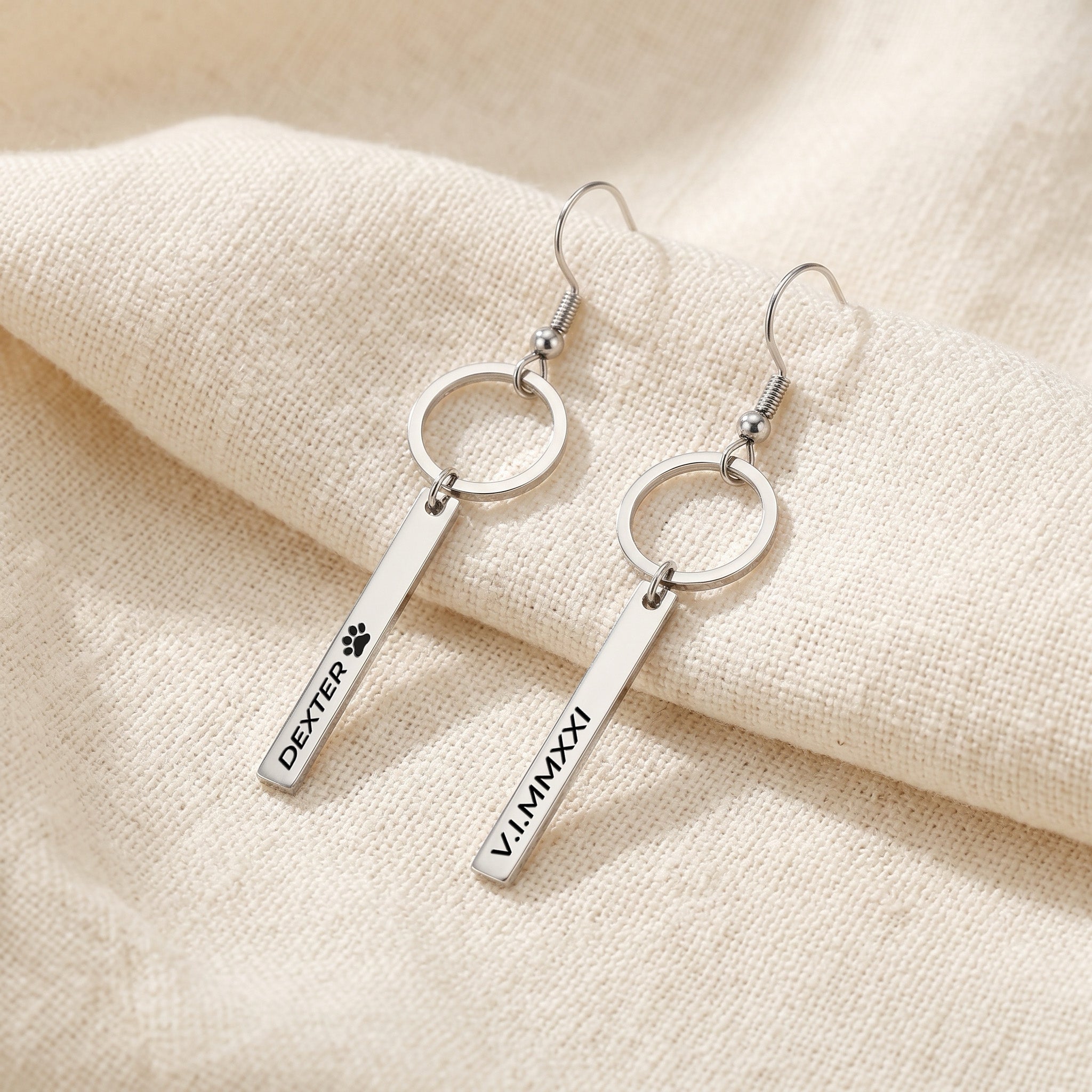 Pair of silver pet name bar drop earrings engraved with a pet's name in block letters, on cream fabric