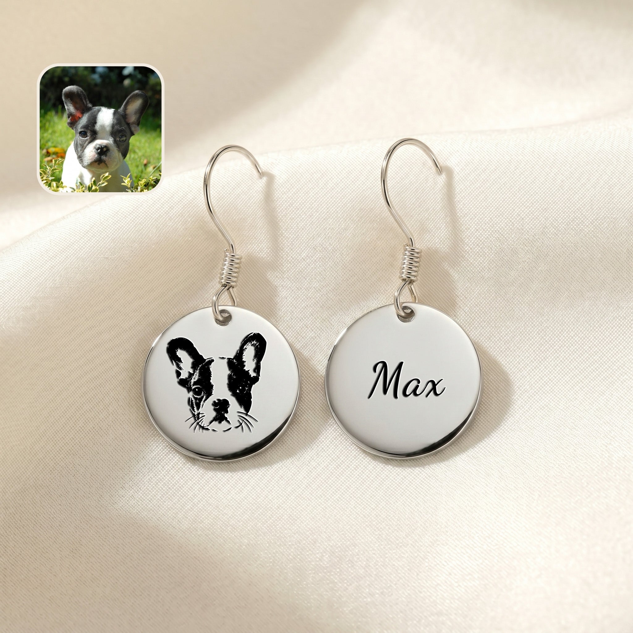 Pair of silver drop earrings with hand-drawn portrait and name discs, staged on cream fabric beside a dog photo