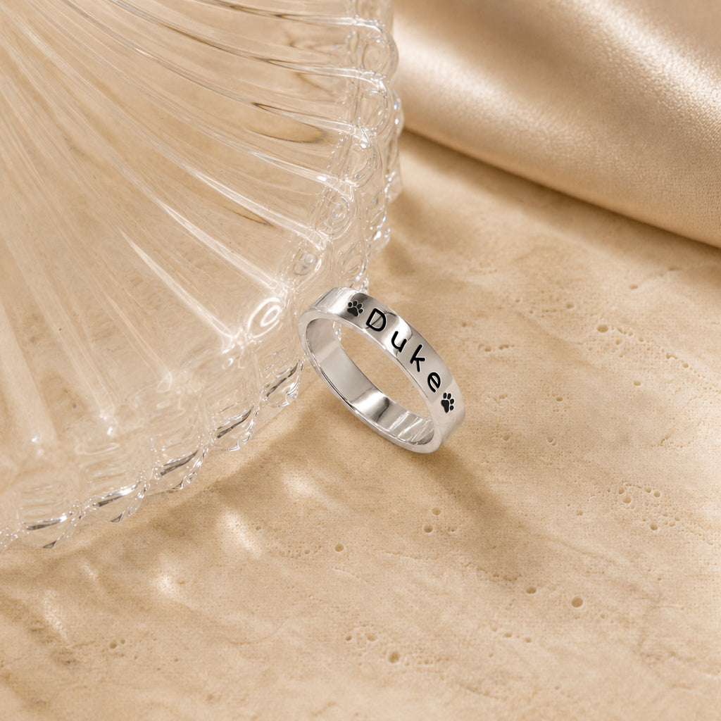 Rhodium-plated silver-tone pet name stacking ring engraved with a name and paw prints, beside a fluted glass dish on travertine