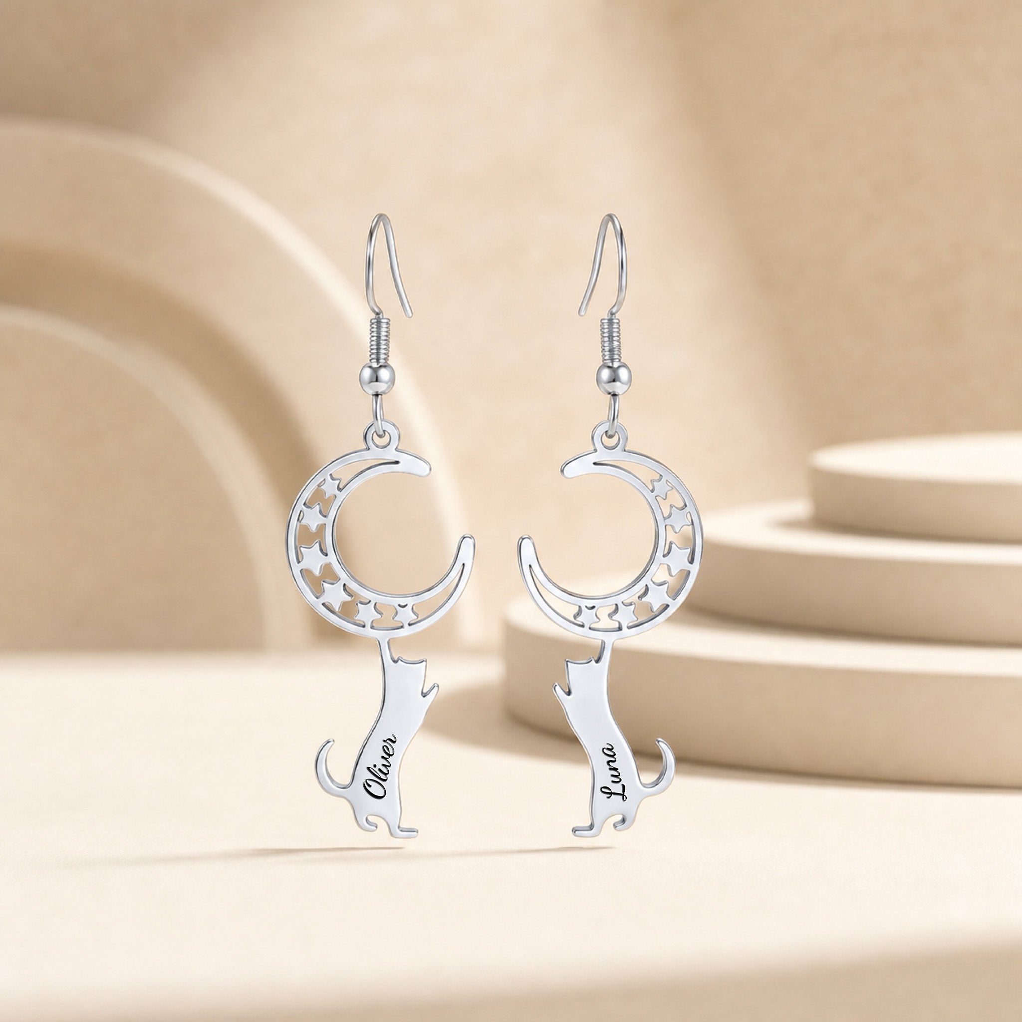 Pair of silver crescent-moon and cat drop earrings with an engraved name, staged upright on a soft beige pedestal