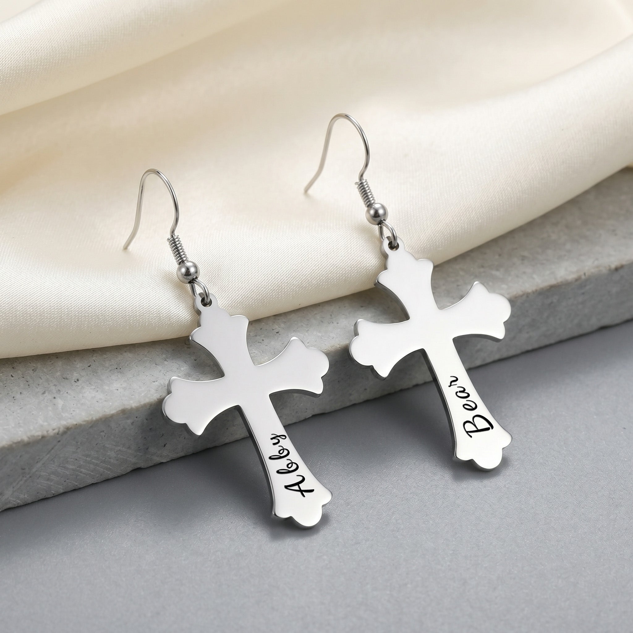 Rhodium silver pet cross earrings shown as a matched pair on a gray stone slab — pet name laser-engraved in script on the lower stem of each cross