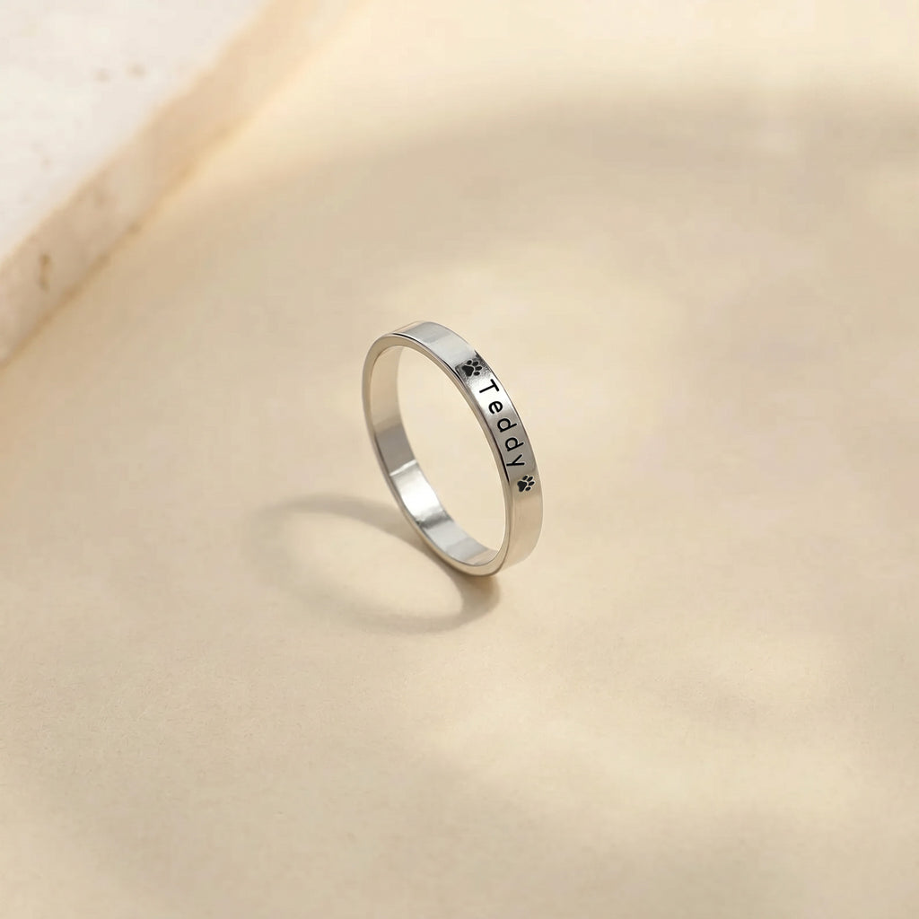 Silver pet name band ring standing on a warm stone surface, name and paw engraving facing forward
