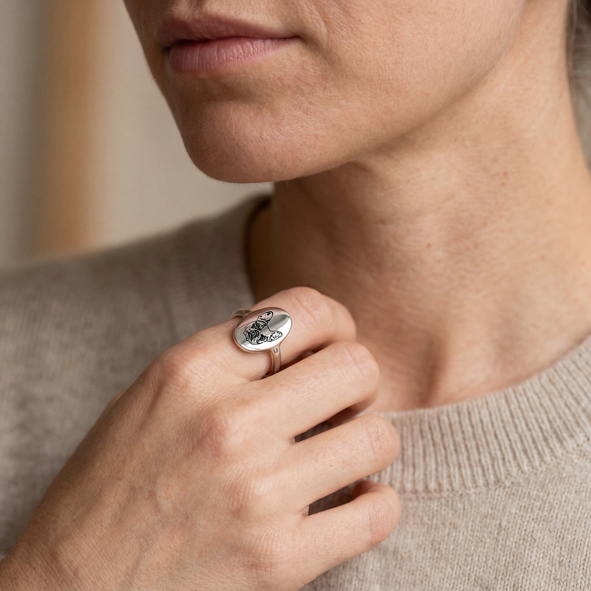Woman wearing a silver oval signet pet portrait ring, hand near her collarbone in a soft neutral knit, indoor close-up