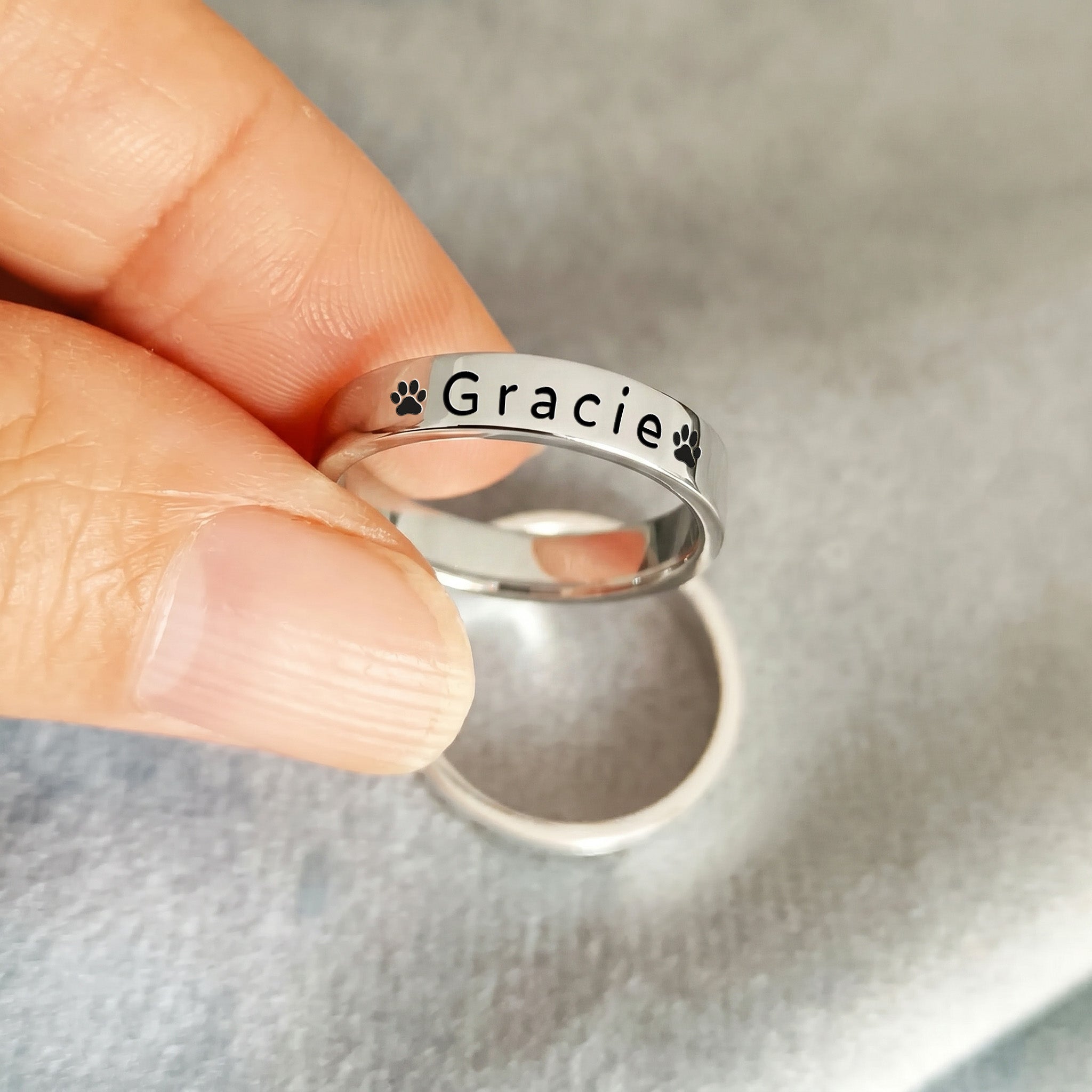 Rhodium-plated pet name stacking ring held between fingertips against a soft grey background