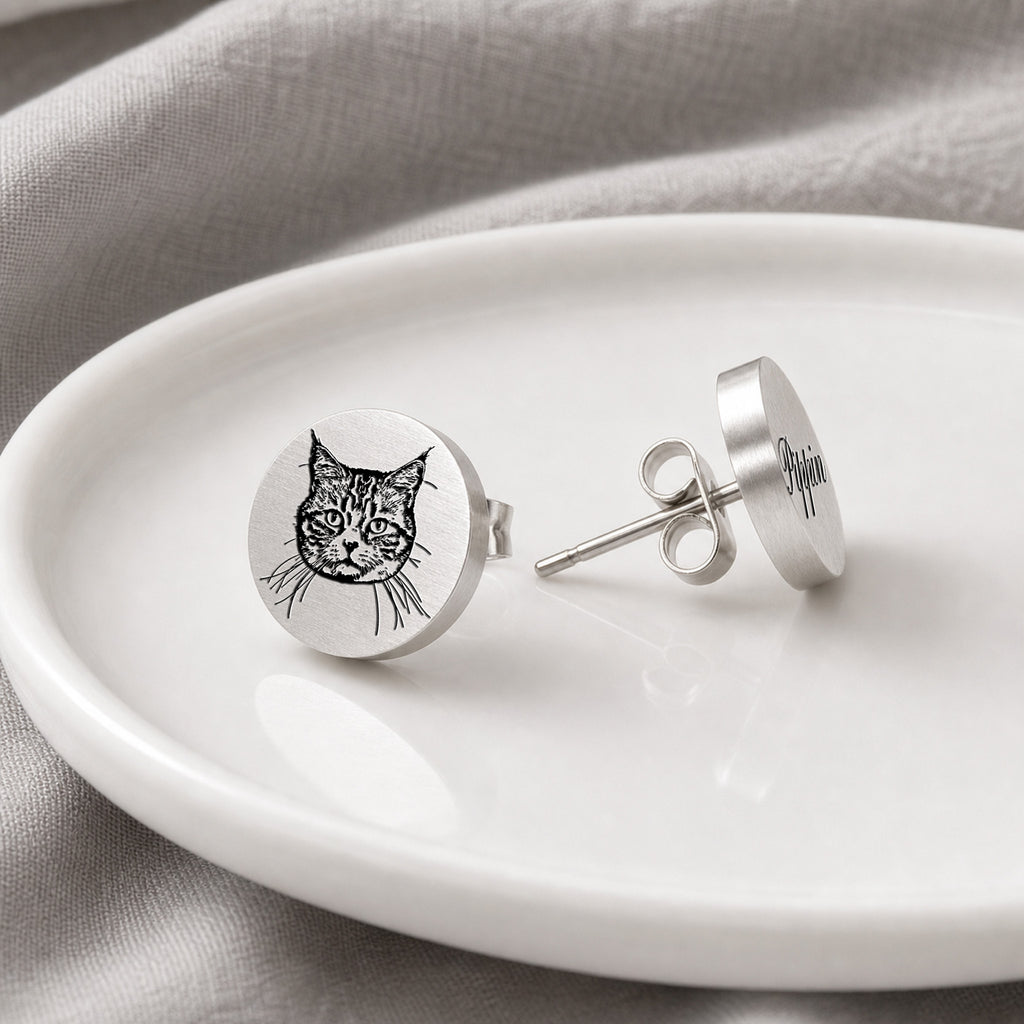Pair of silver pet portrait stud earrings on a white dish, portrait stud beside engraved name stud