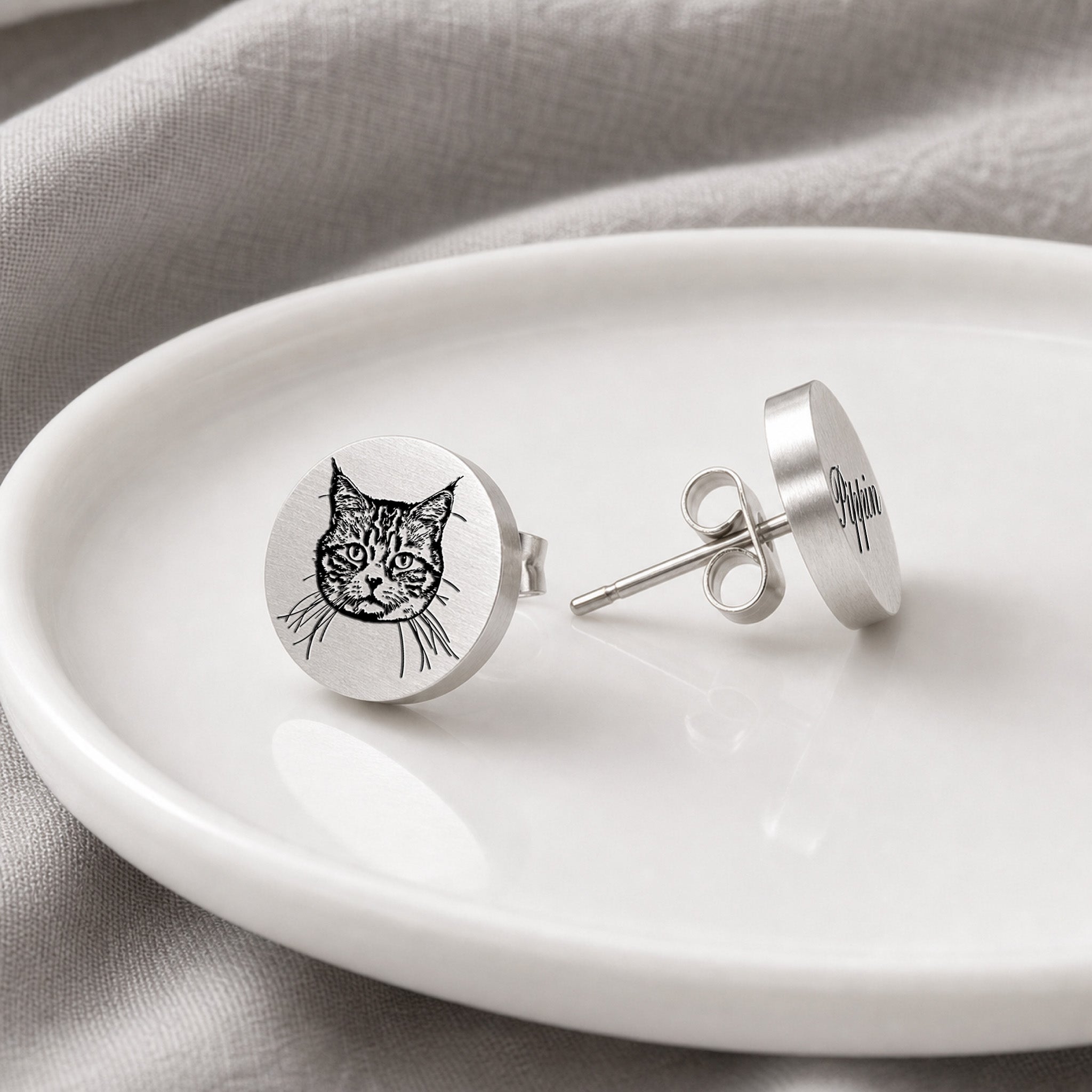 Pair of silver pet portrait stud earrings on a white dish, portrait stud beside engraved name stud