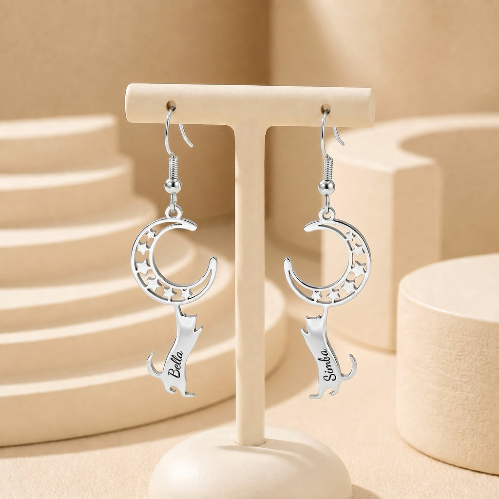 Silver cat-and-moon name earrings hanging from a cream display stand in a neutral ceramic tabletop scene