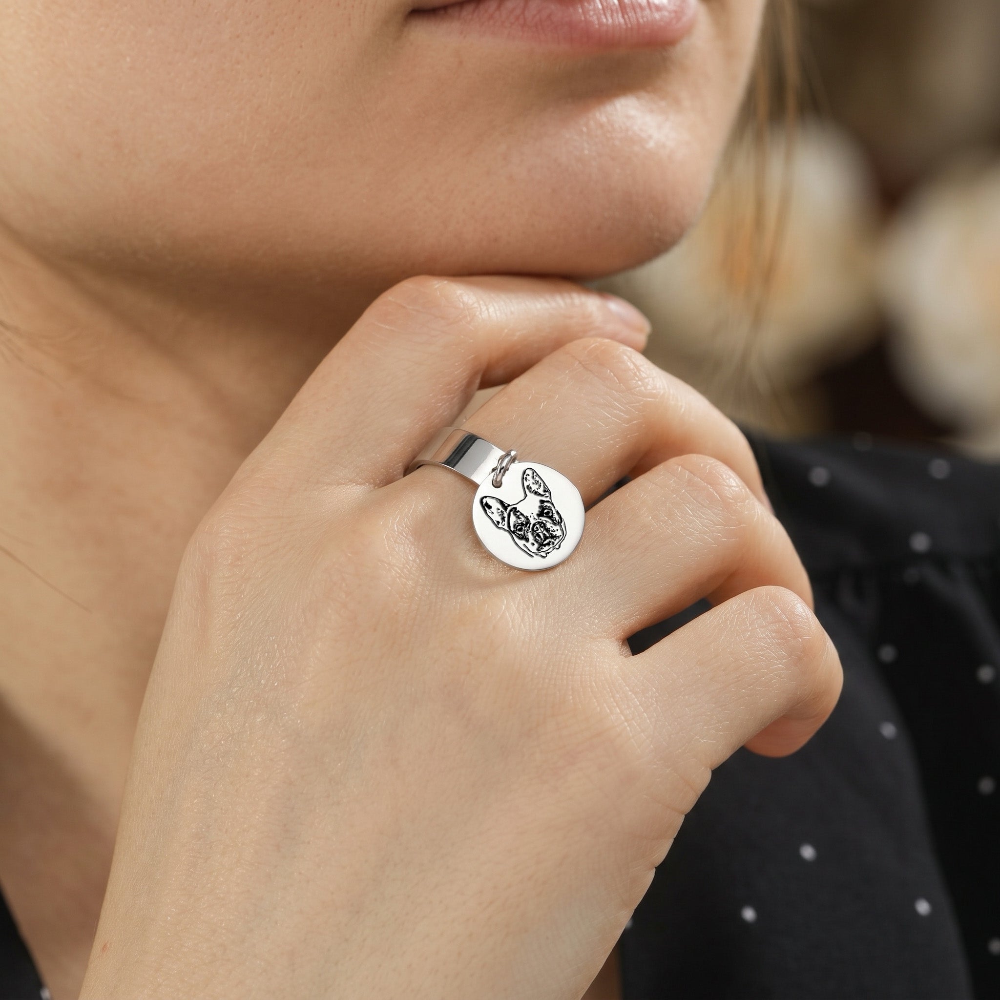 Silver-tone pet portrait disc ring worn on a finger, held up beside the wearer's face indoors