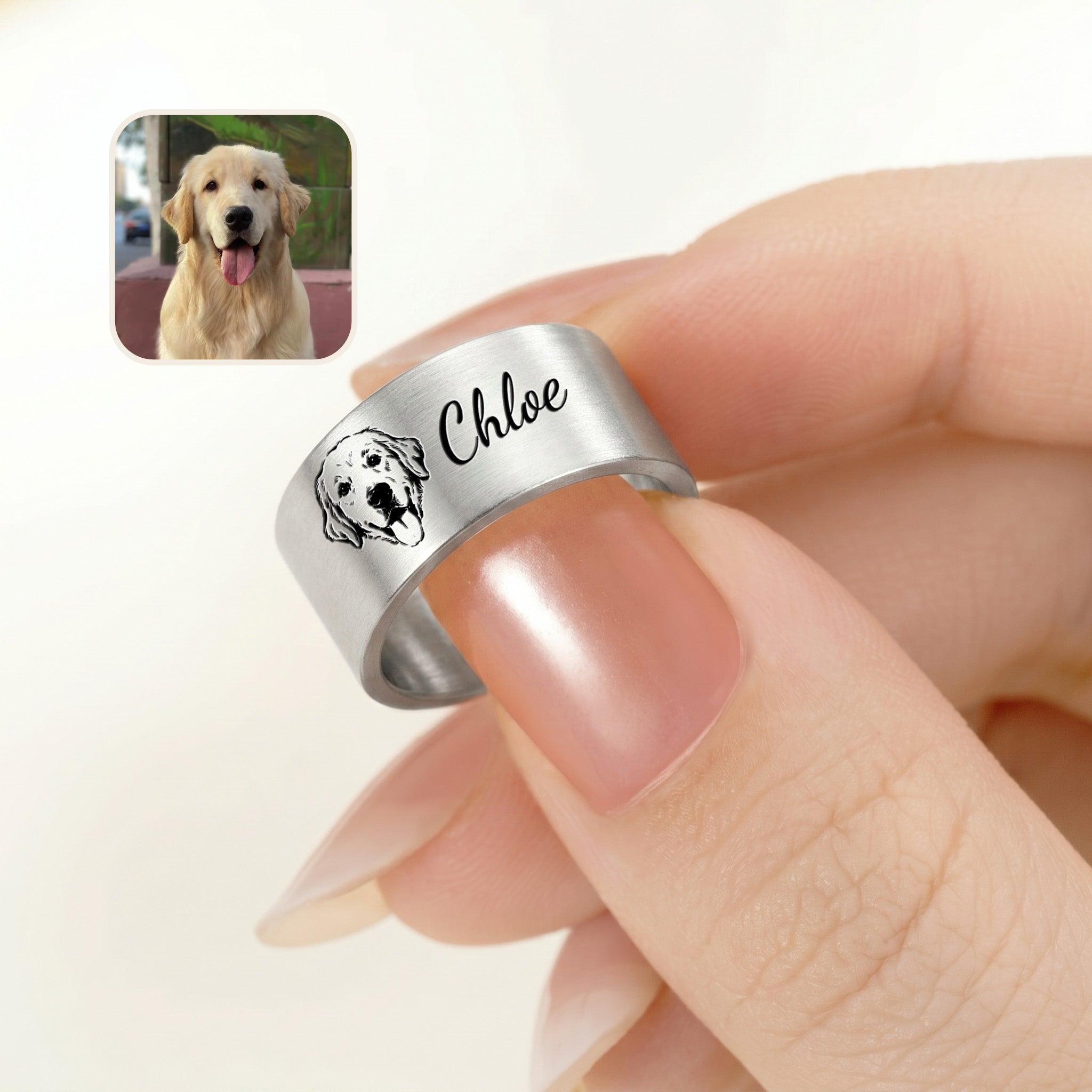 Brushed silver wide band pet ring pinched between fingers, its engraved portrait and script name in view, beside a golden retriever photo