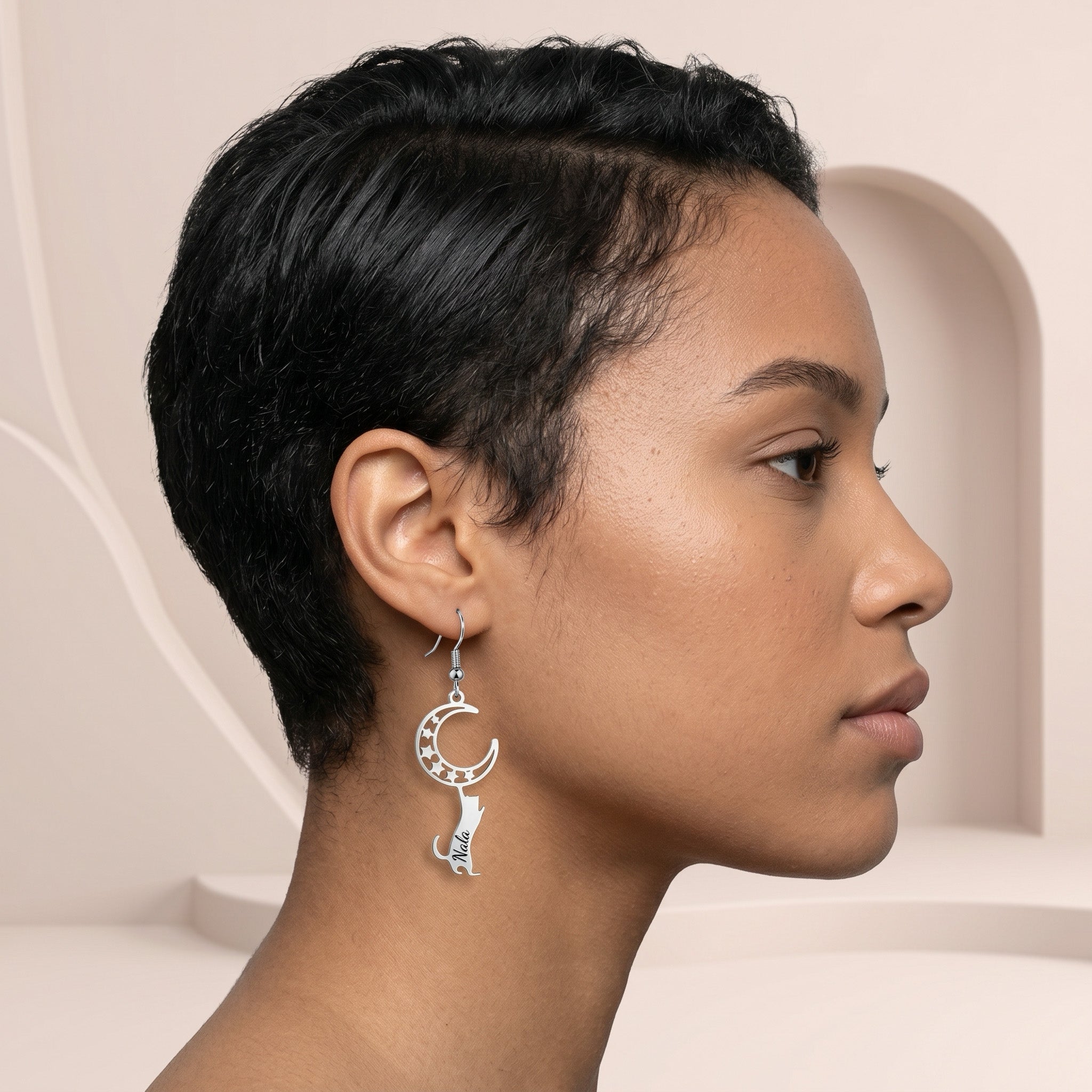 Silver cat-and-moon name earring worn in side profile against a blush-beige sculptural backdrop
