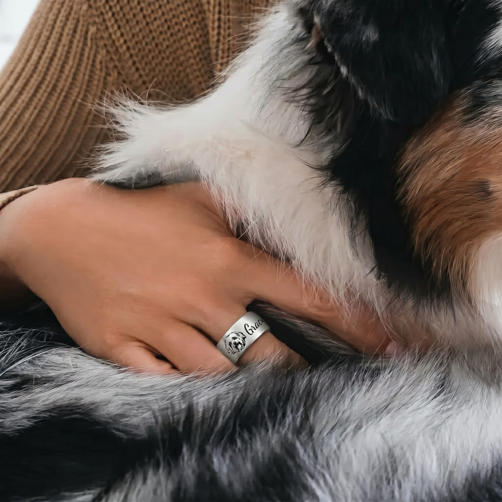 Silver wide band pet ring worn on a finger, engraved portrait and script name, a hand resting near a dog in a home scene
