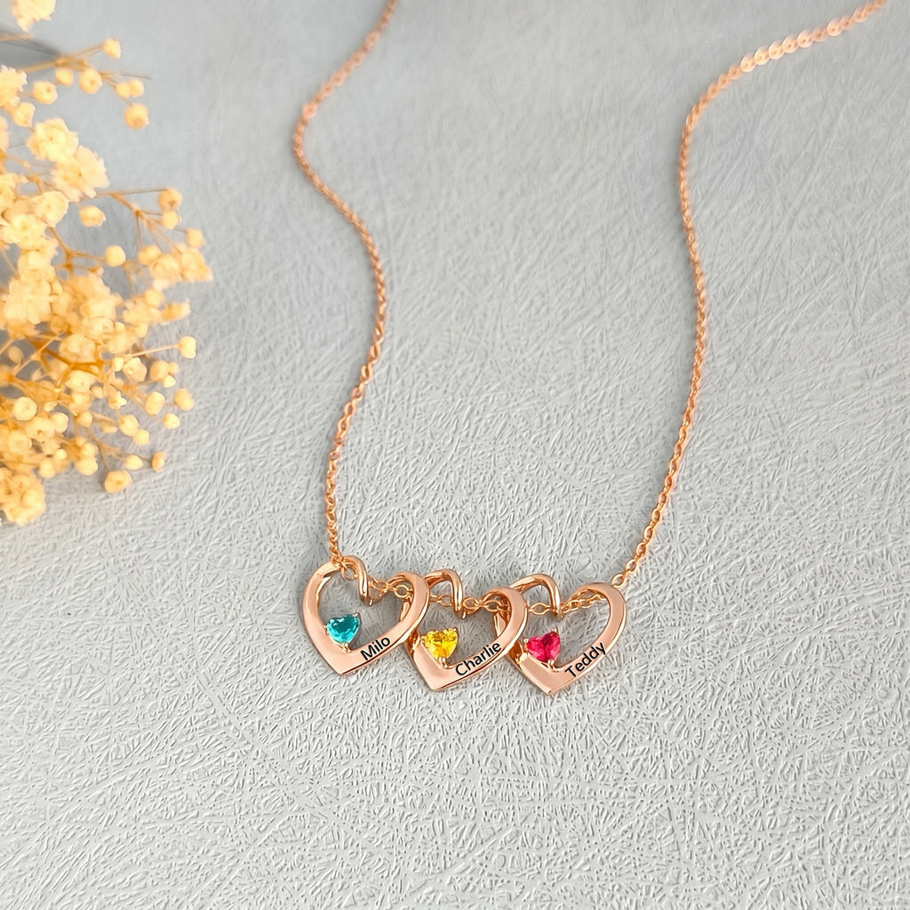 Three rose gold heart pendants with colorful birthstones and pet names on cream background